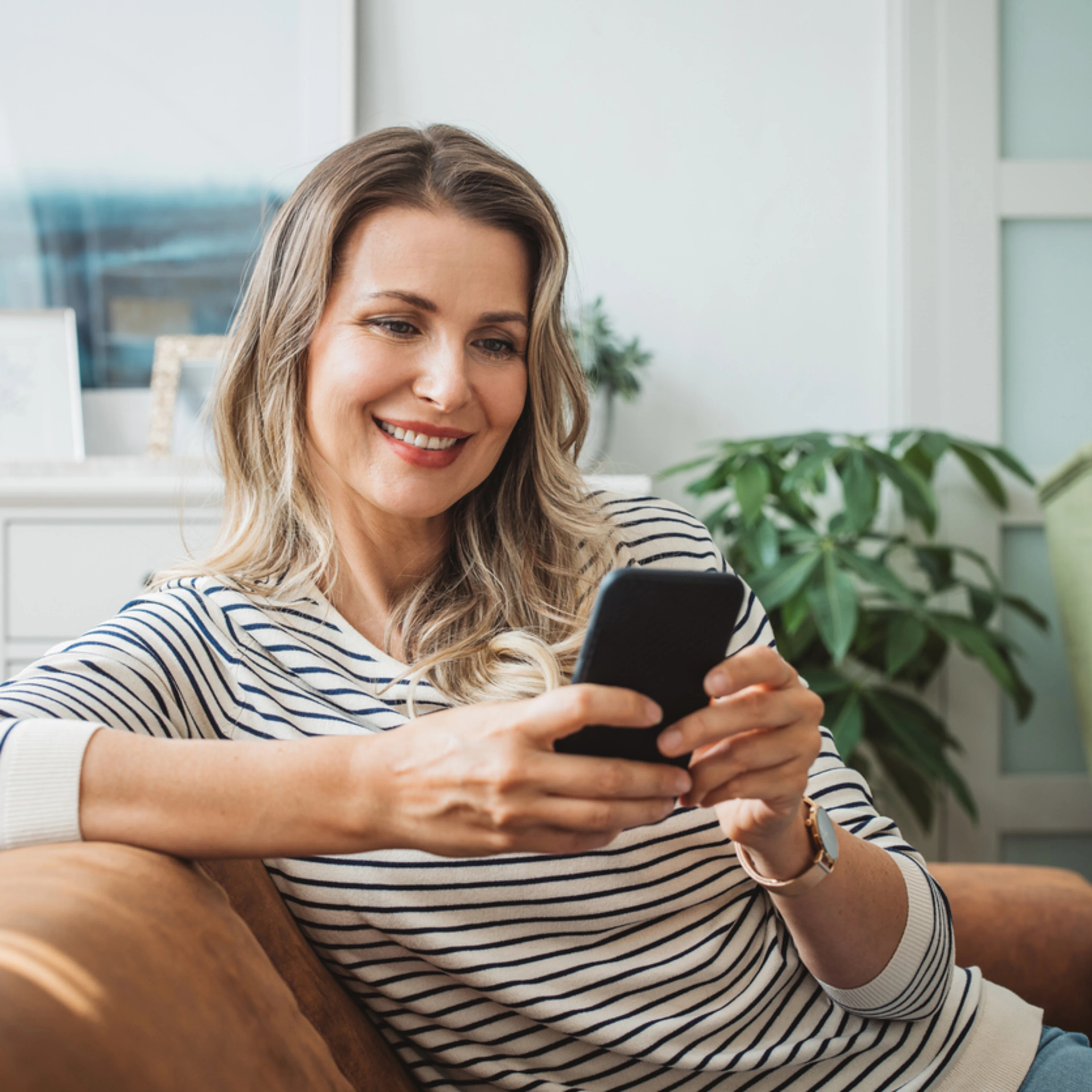 Smiling woman in striped shirt sits on a couch, looking at her smartphone. The room is cozy, with plants and framed art, creating a warm, relaxed atmosphere.