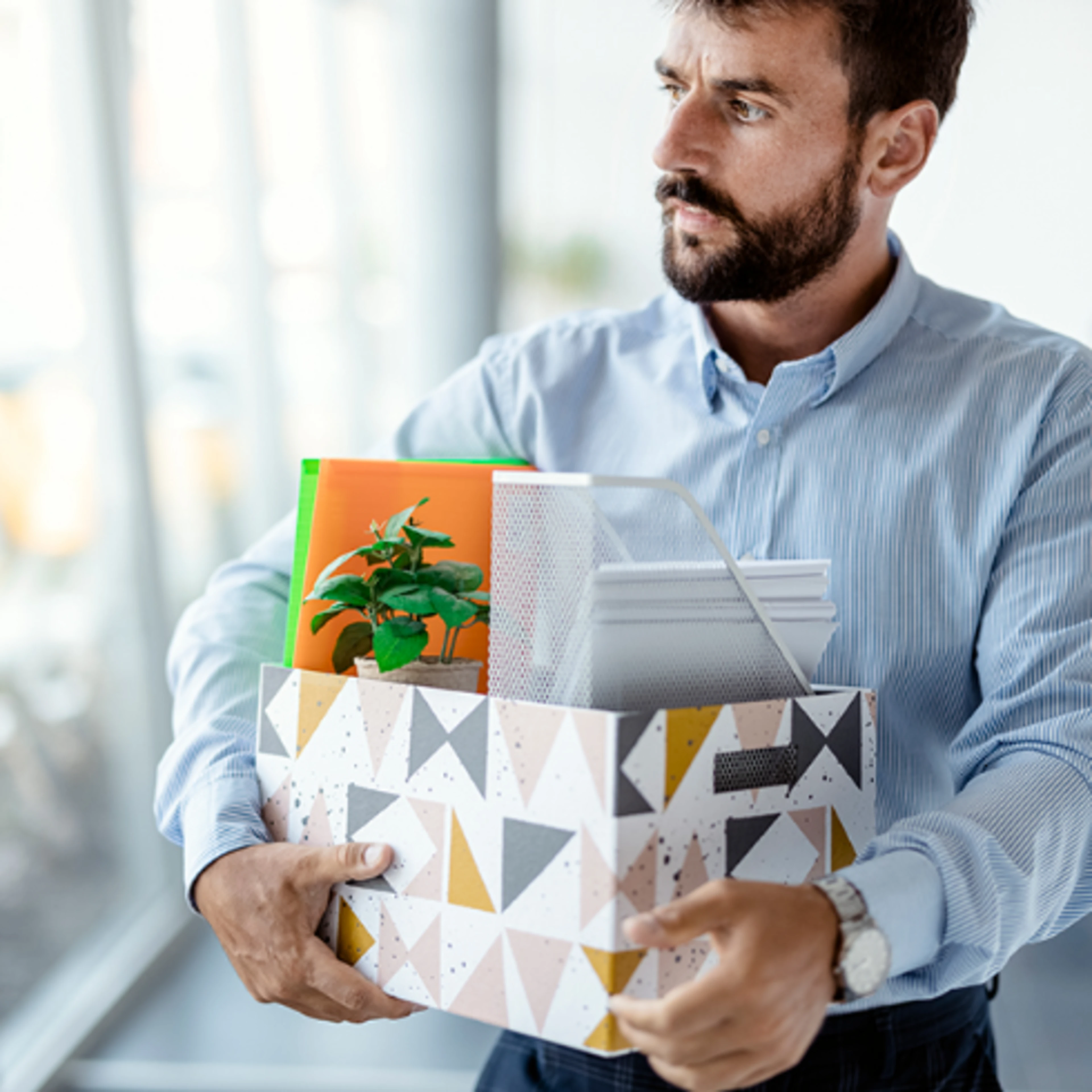 A man with a serious expression holds a decorative box containing office supplies and a small plant, walking in a bright office with blurred colleagues in the background.