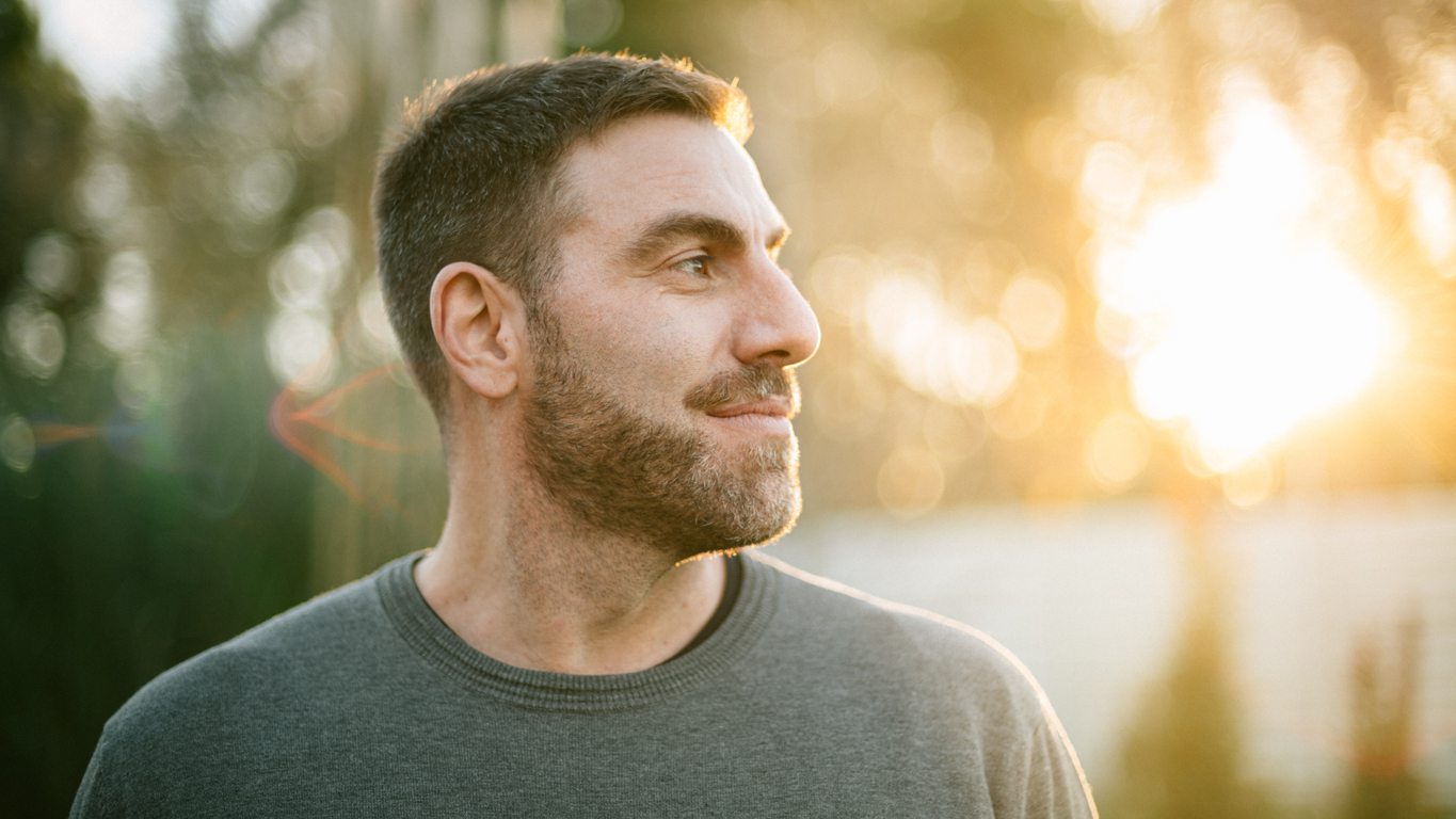 A man with a beard smiles slightly, looking to the right, in a sunlit garden. The warm evening sunlight creates a peaceful, contemplative mood.