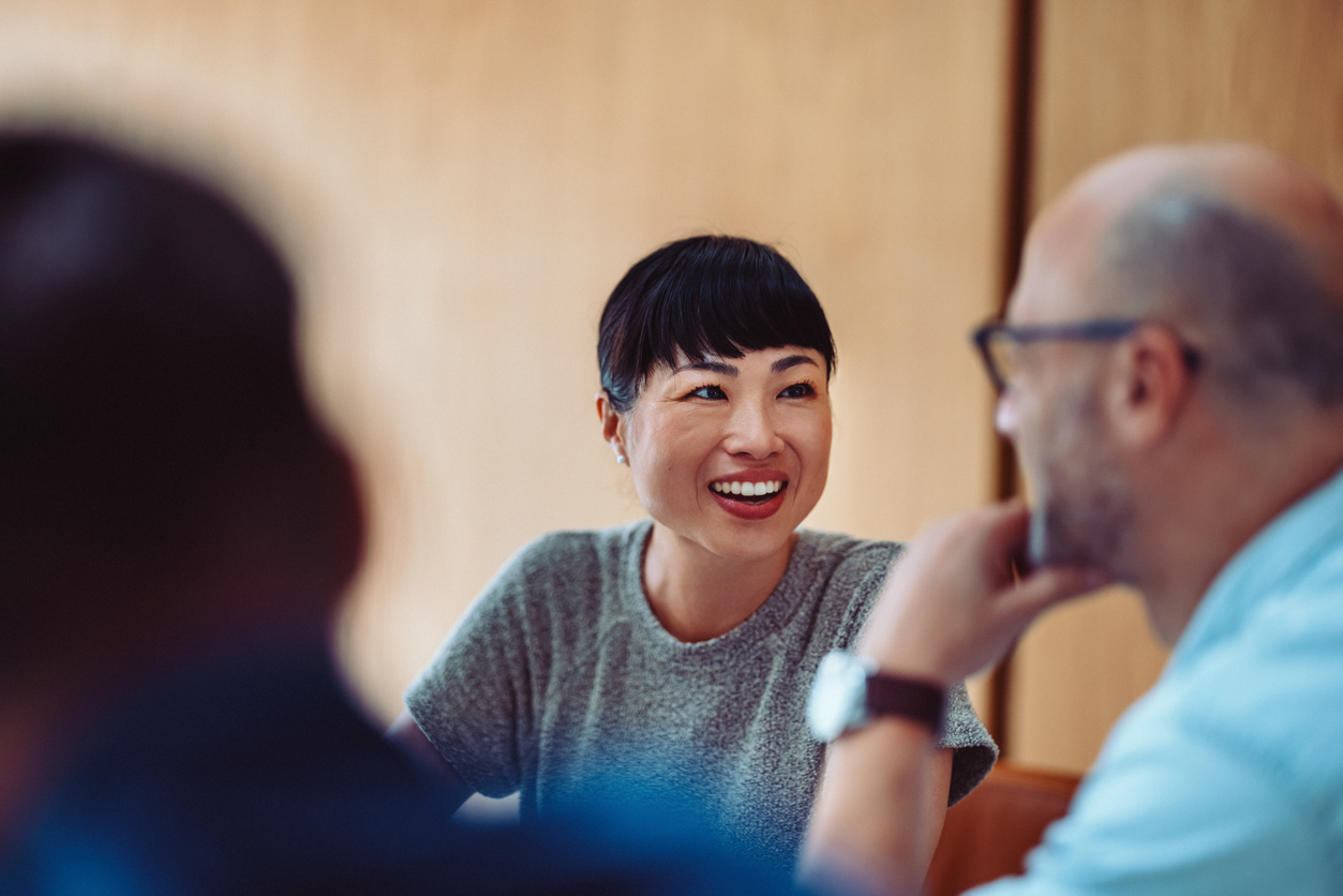 A smiling woman with short black hair chats with colleagues in a modern office. She wears a gray t-shirt while a man in a light blue shirt listens. Wood-paneled walls and natural lighting create a collaborative atmosphere.