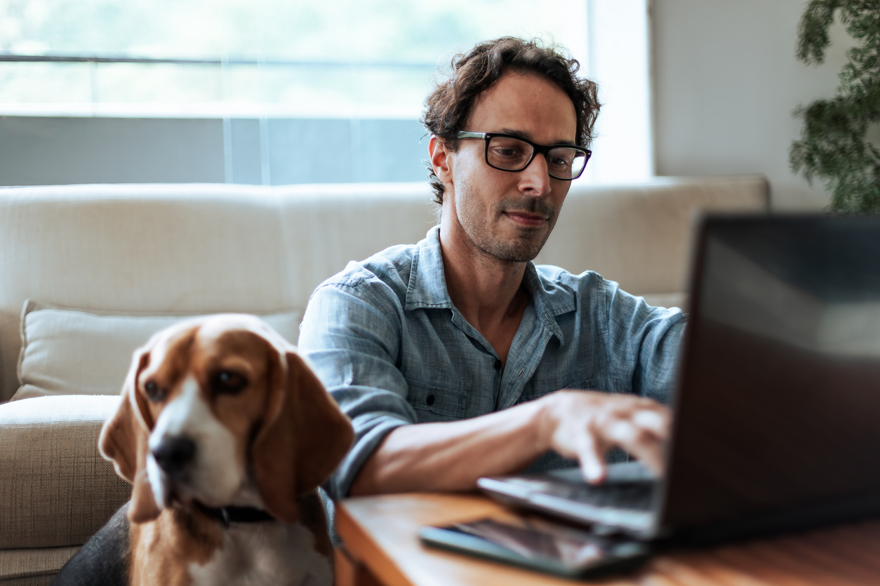 A man is focused on his laptop while sitting on a couch. He wears glasses and a casual shirt. Beside him, a calm beagle looks towards the camera.