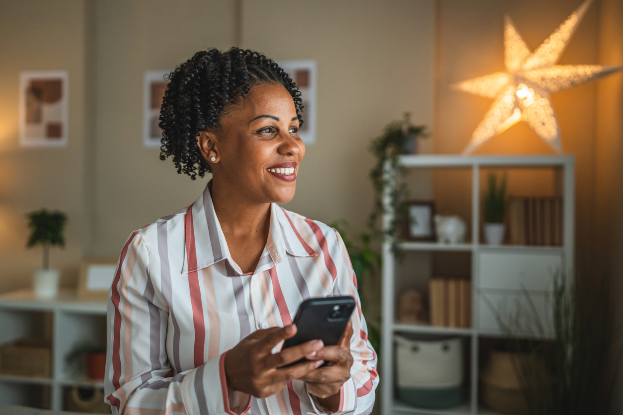 Smiling woman with curly hair holds a smartphone in a cozy, well-lit room. Shelves and a glowing star light create a warm, inviting atmosphere.