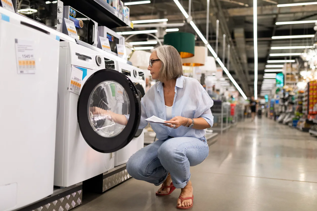 A woman with silver hair and glasses examines a front-loading washing machine in a large retail store. She holds product information while crouching to inspect the appliance, thoughtfully considering a major purchase.
