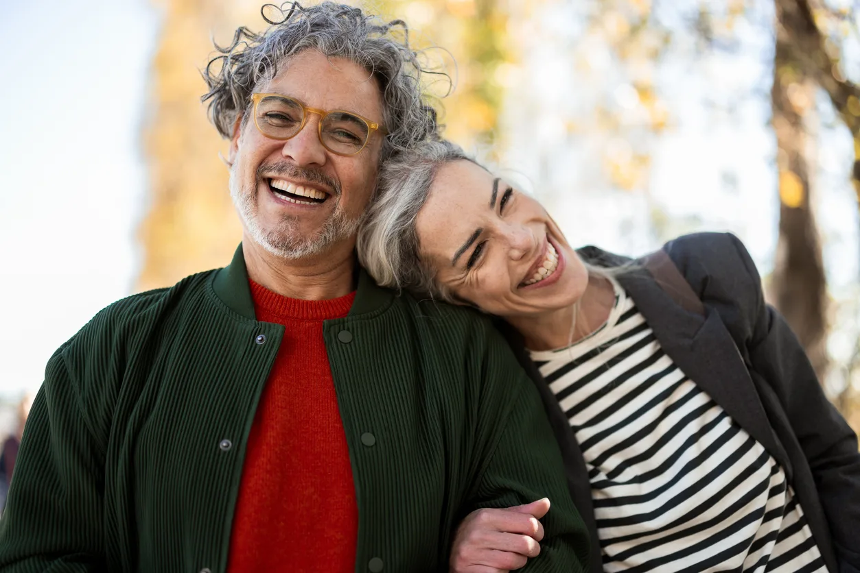 Smiling couple in casual attire embrace outdoors, with autumn trees in soft focus. They display warmth and happiness, leaning close together.