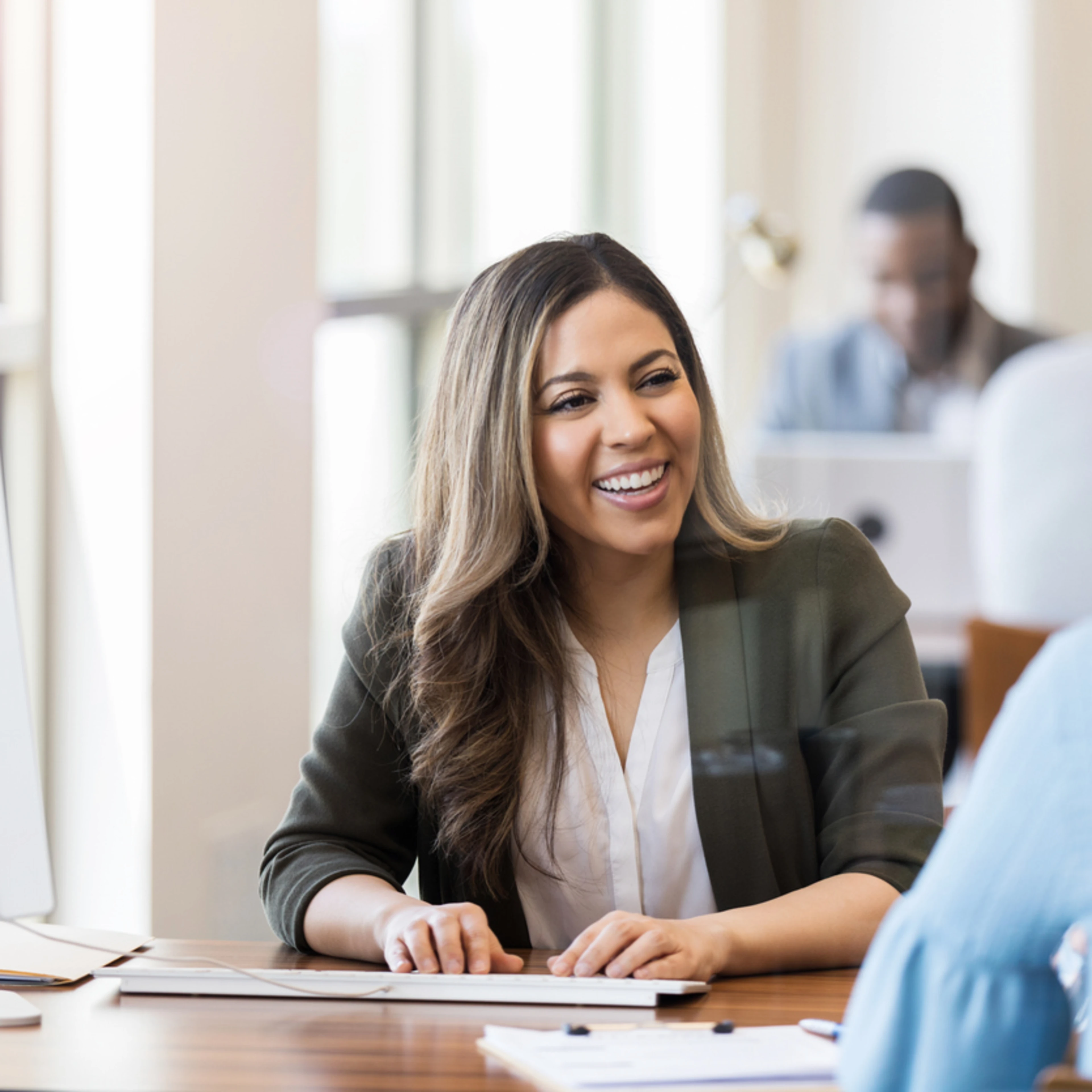 A smiling young woman with long brown hair in an olive blazer provides friendly customer service across a wooden desk. She faces an older woman with short gray hair in a bright, modern office with large windows and natural light.