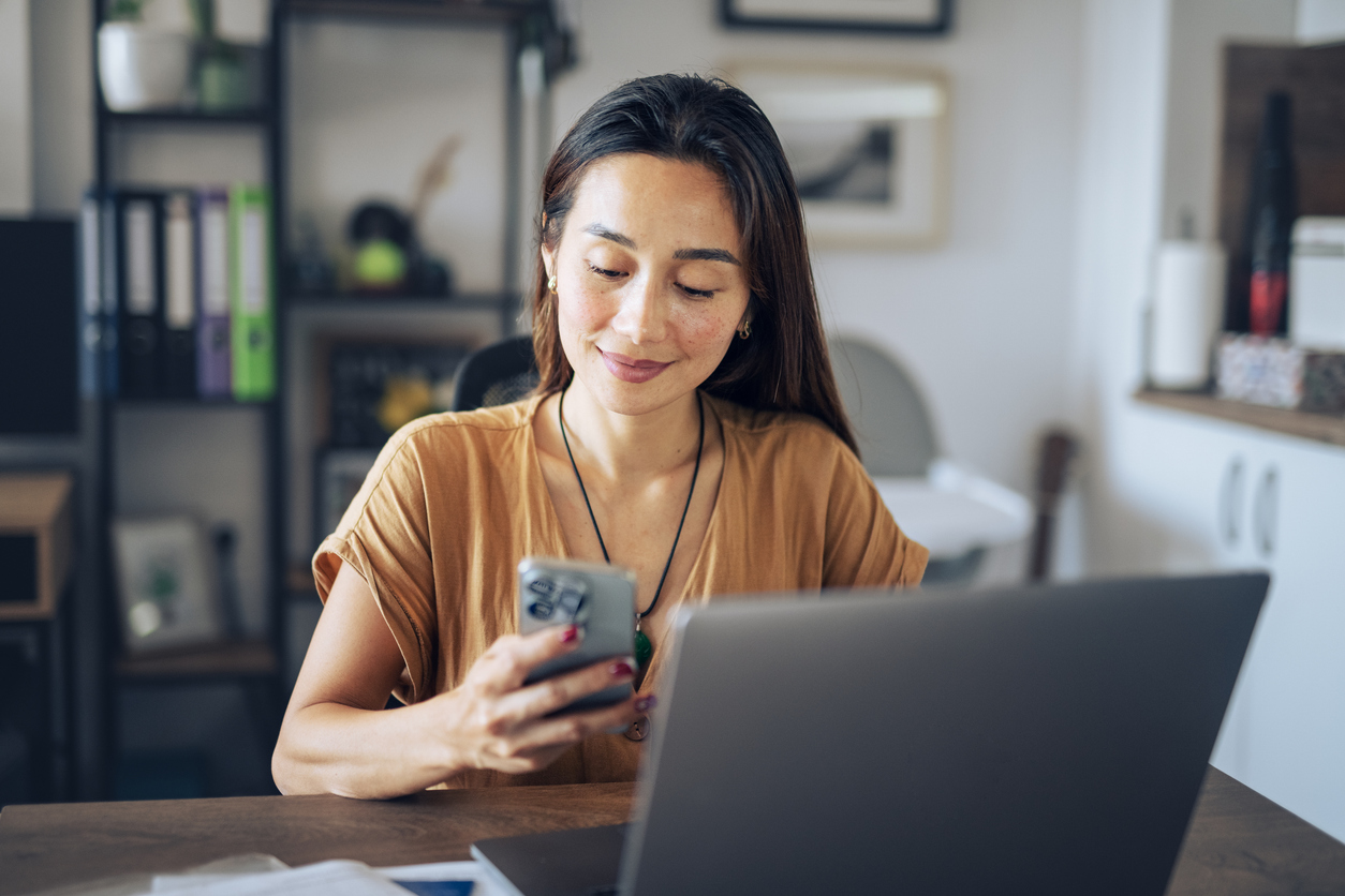 Woman in a cozy office setting, smiling at her phone. A laptop is open on the desk with shelves and decor in the background, conveying productivity.