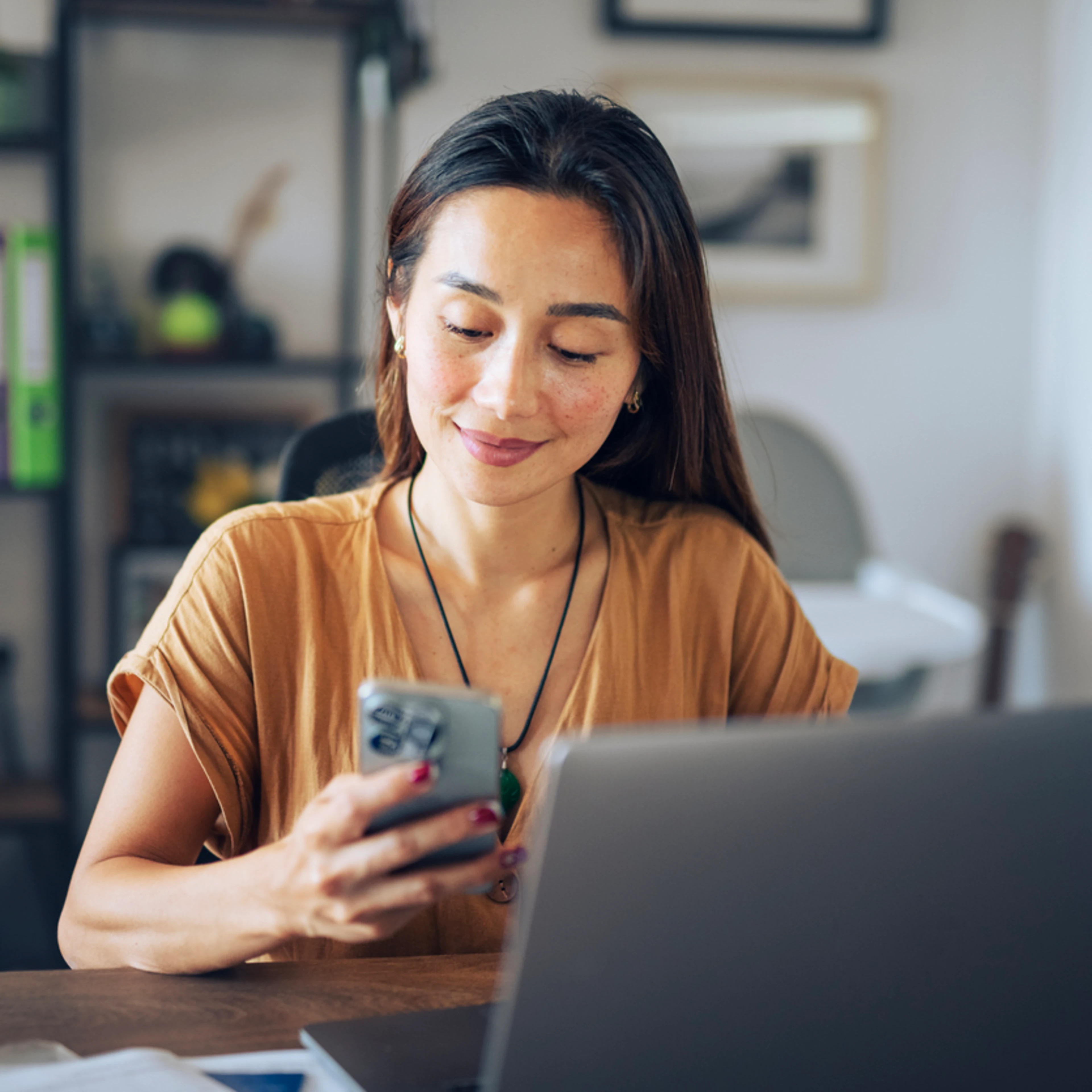 Woman in a cozy office setting, smiling at her phone. A laptop is open on the desk with shelves and decor in the background, conveying productivity.