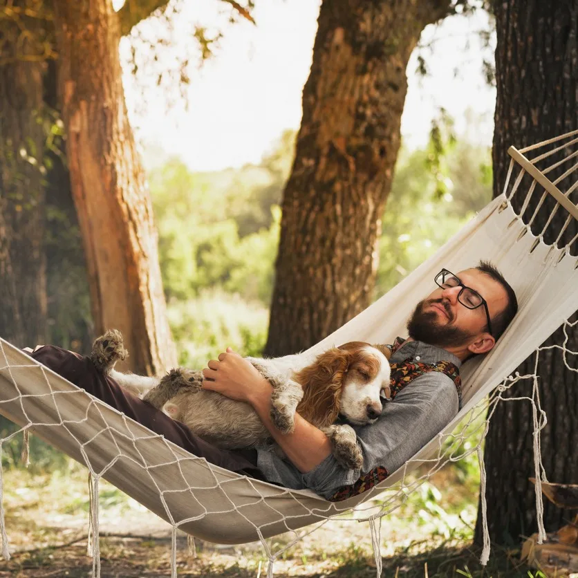 A man relaxes in a hammock under trees, gently holding a sleeping dog. Sunlight filters through leaves, creating a peaceful, serene ambiance.