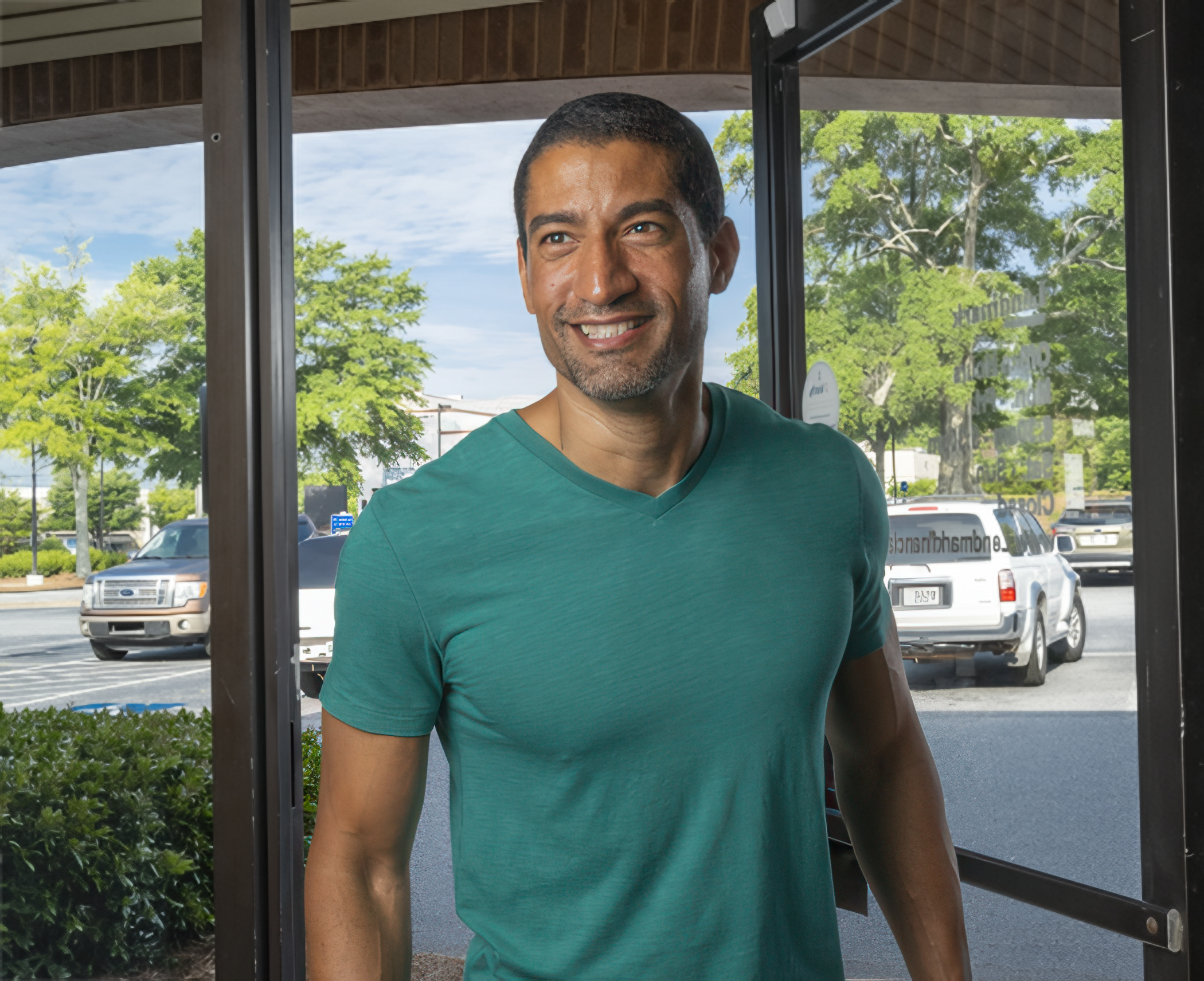 A man wearing a teal T-shirt smiles warmly while entering a building. Glass doors and parked cars outside suggest a casual, sunny day.
