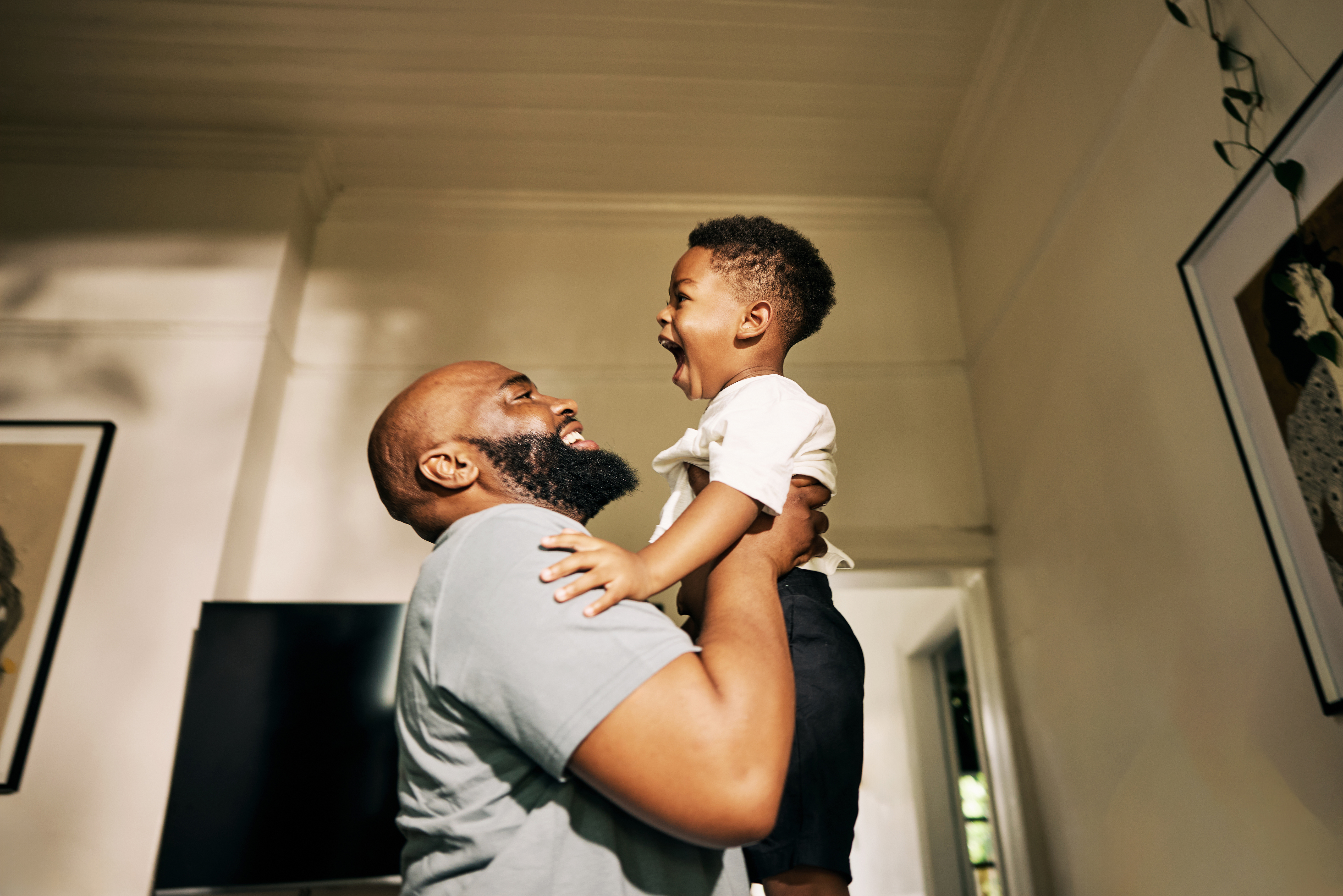 A joyful moment as a father lifts his laughing young son in a sunlit room, creating an atmosphere of warmth and happiness. Family bond is evident.