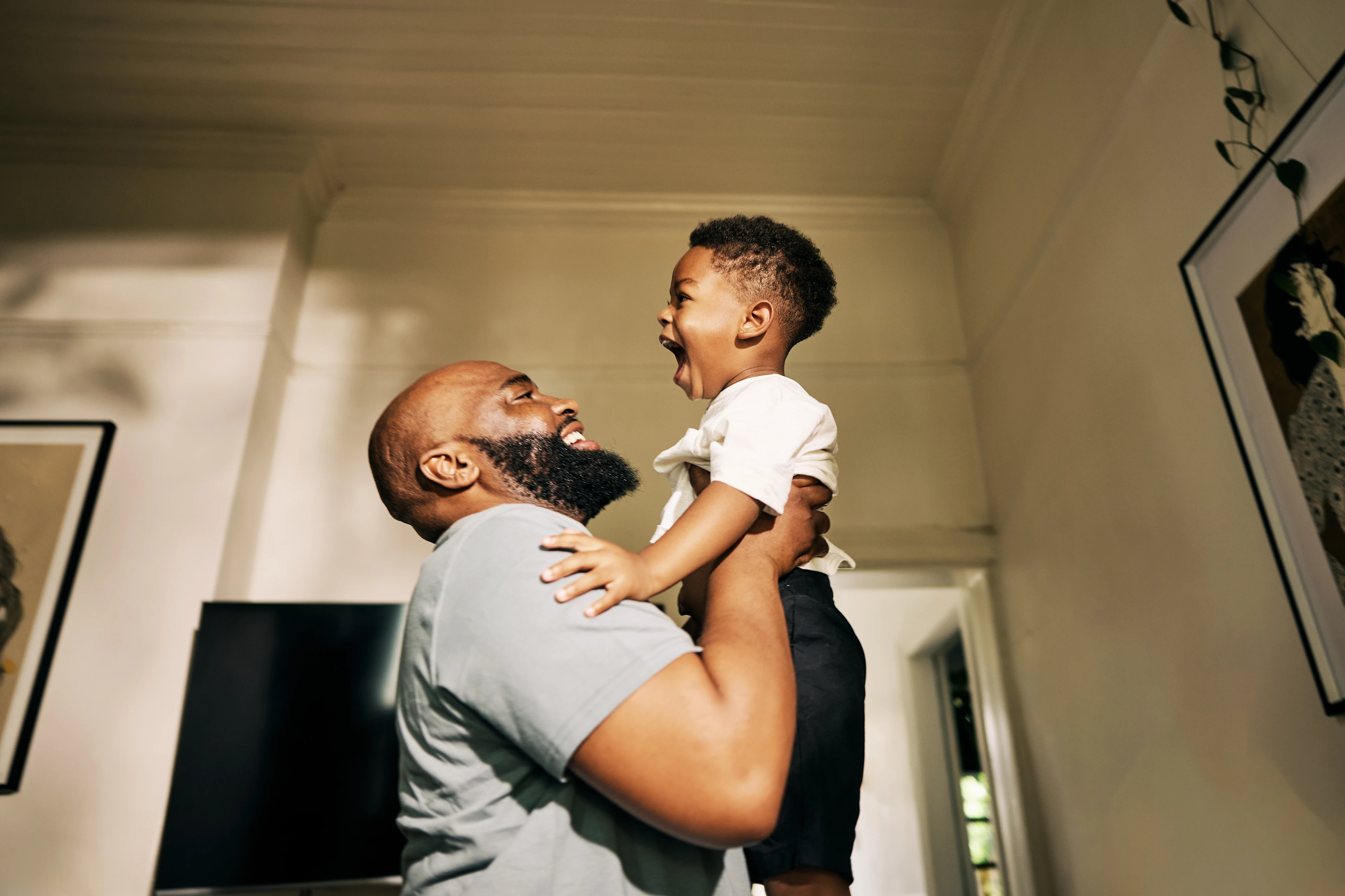 A joyful moment as a father lifts his laughing young son in a sunlit room, creating an atmosphere of warmth and happiness. Family bond is evident.