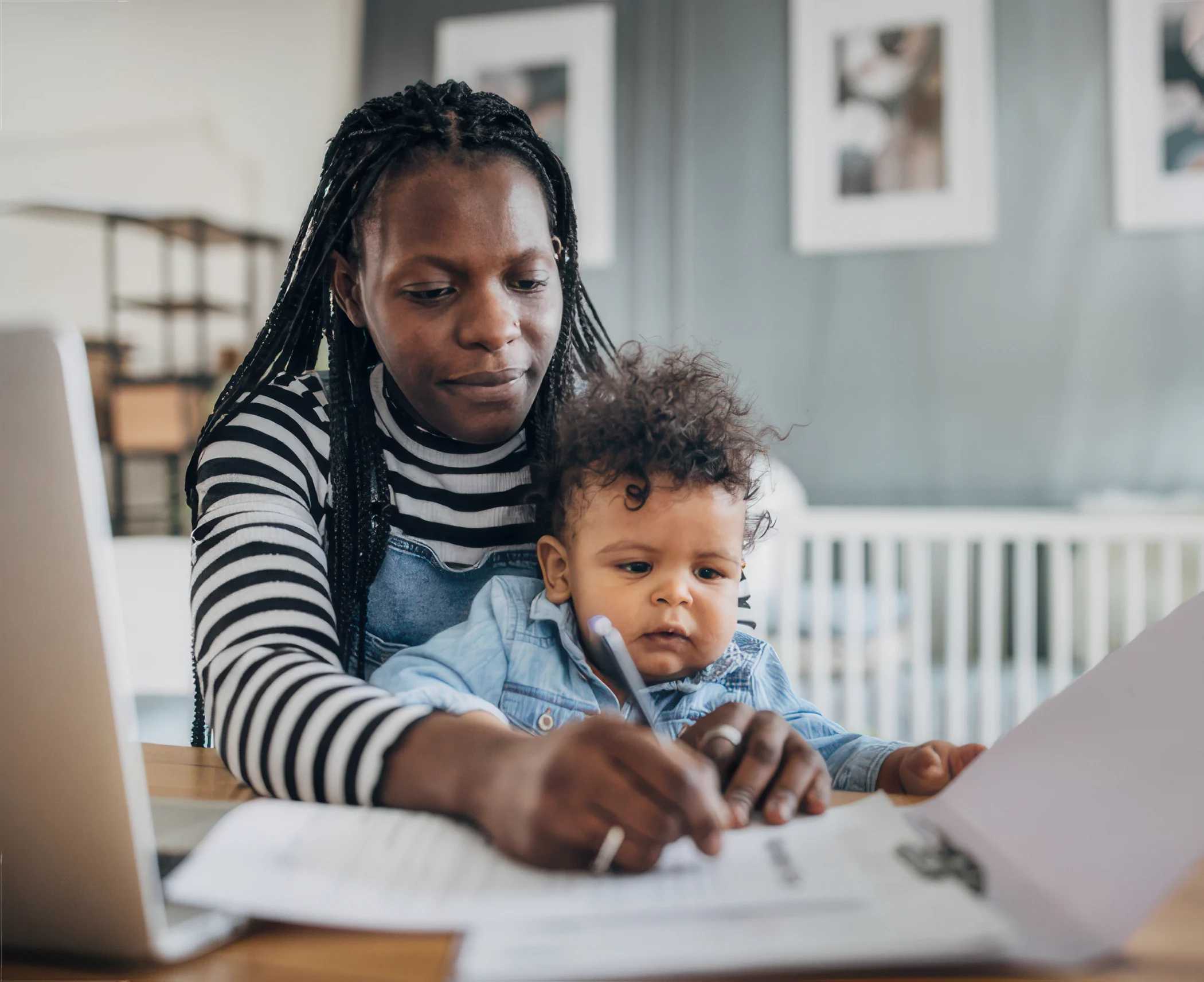 A woman with braided hair focuses on paperwork at a table, holding a pen, with a toddler on her lap. The setting is a cozy, bright home.
