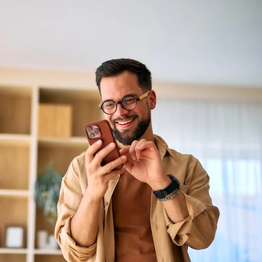 A bearded man wearing tortoiseshell glasses and a tan shirt smiles while using his smartphone in a bright, modern living space. Built-in shelving with plants and books creates a warm, minimalist backdrop.