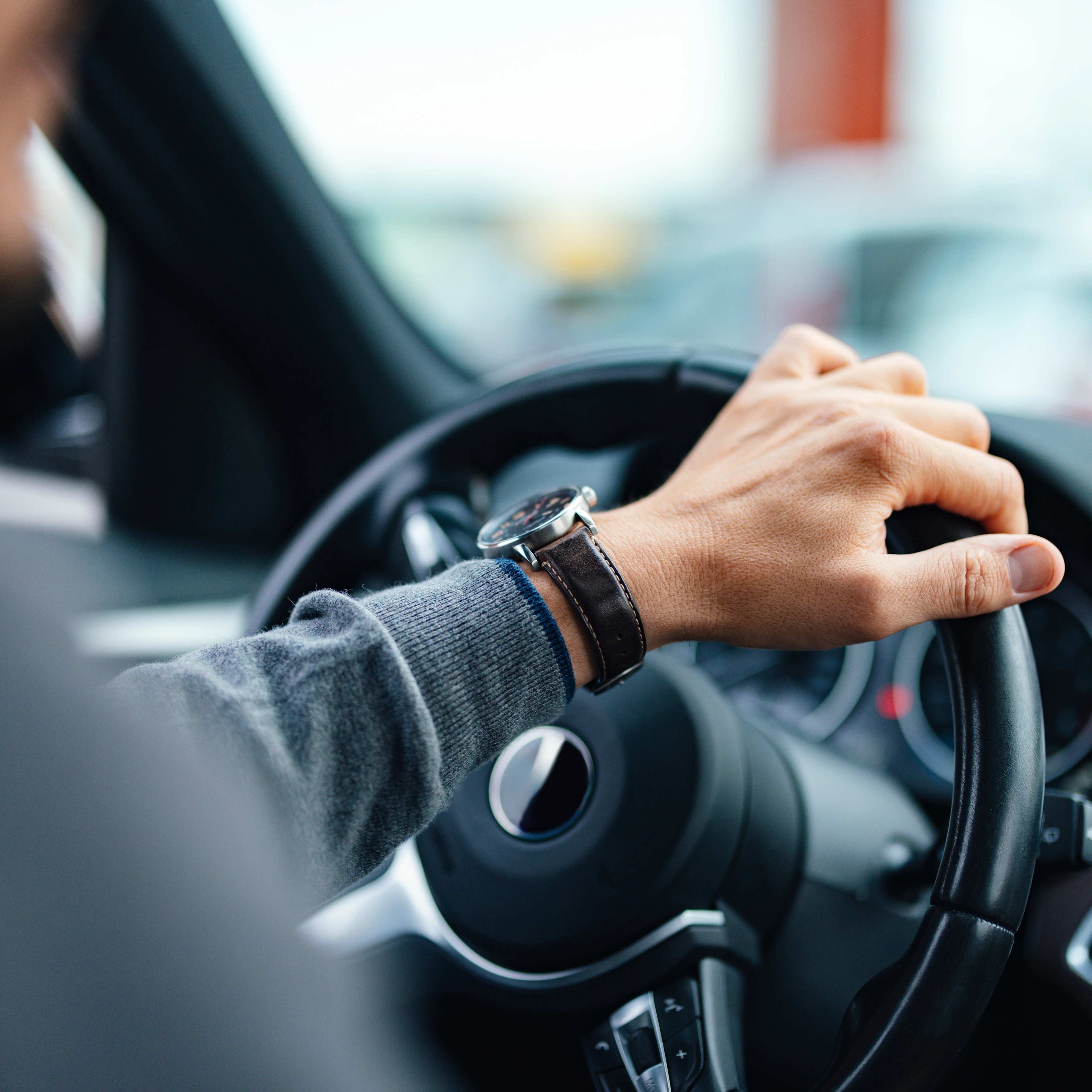 Close-up of a person’s hand wearing a watch on a steering wheel, implying focus and control while driving. Blurred urban background.