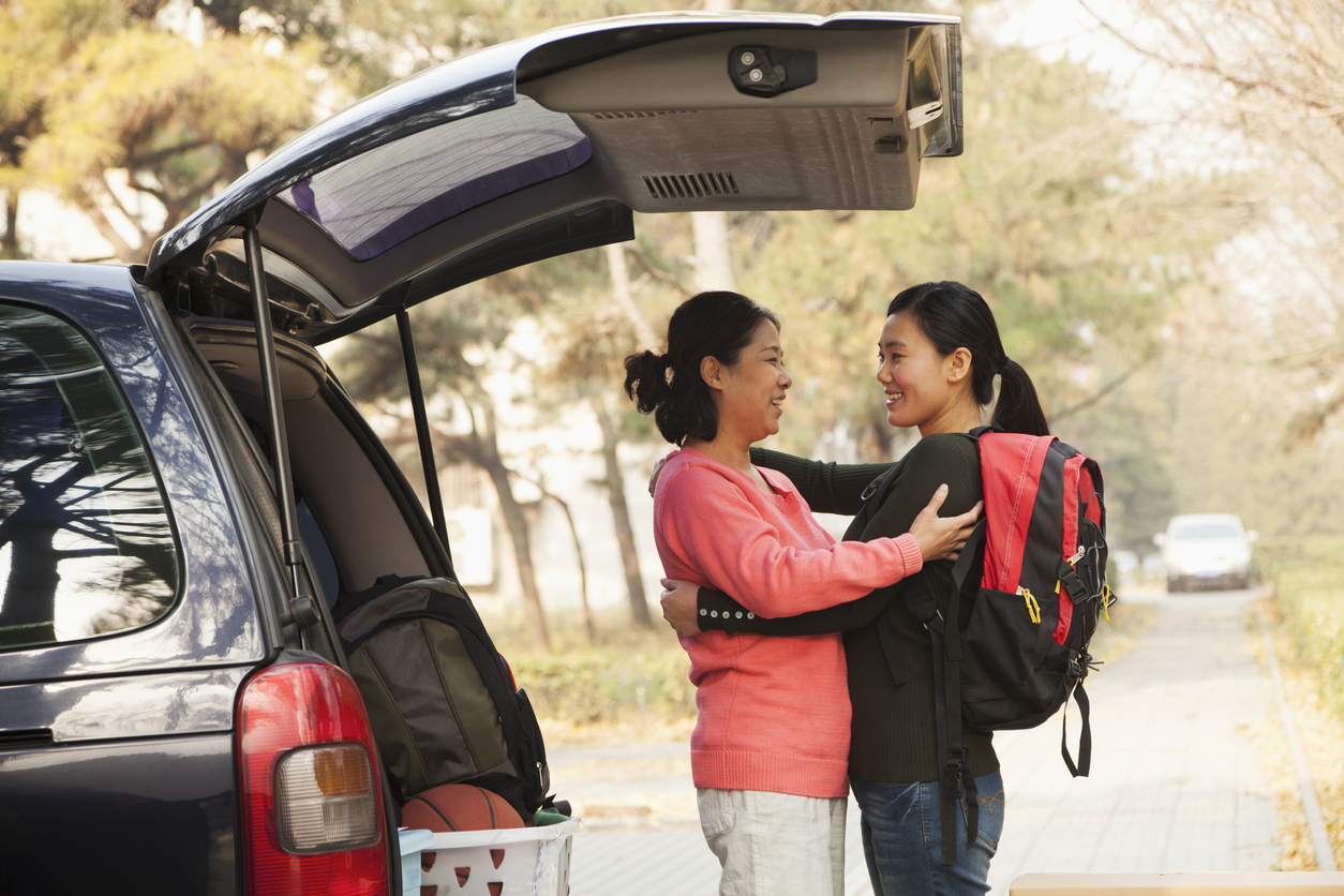 An Asian mother and daughter embrace beside a car trunk loaded with college move-in supplies. The mother wears a coral pink sweater while the daughter carries a backpack. Golden afternoon light captures this bittersweet farewell moment.