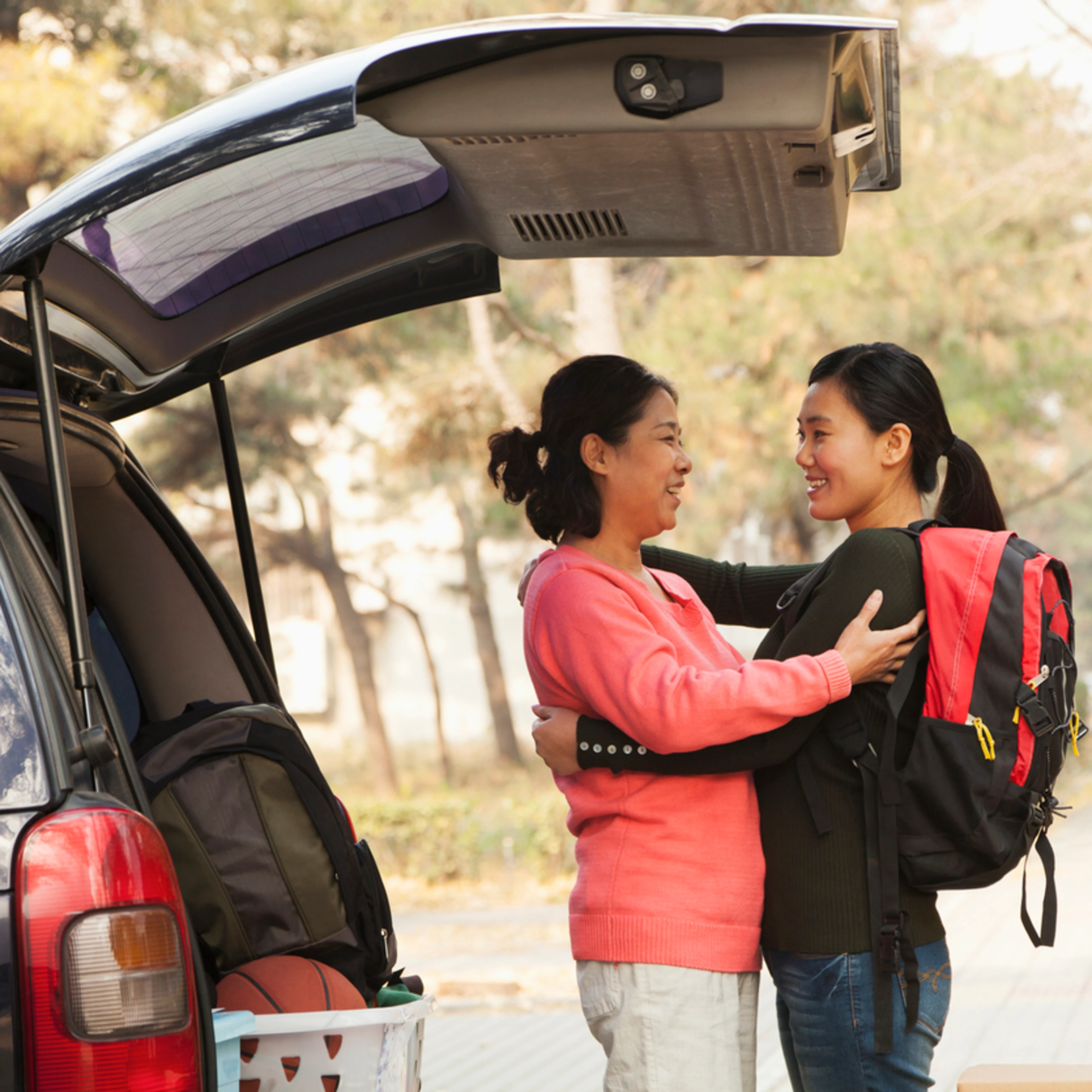 An Asian mother and daughter embrace beside a car trunk loaded with college move-in supplies. The mother wears a coral pink sweater while the daughter carries a backpack. Golden afternoon light captures this bittersweet farewell moment.
