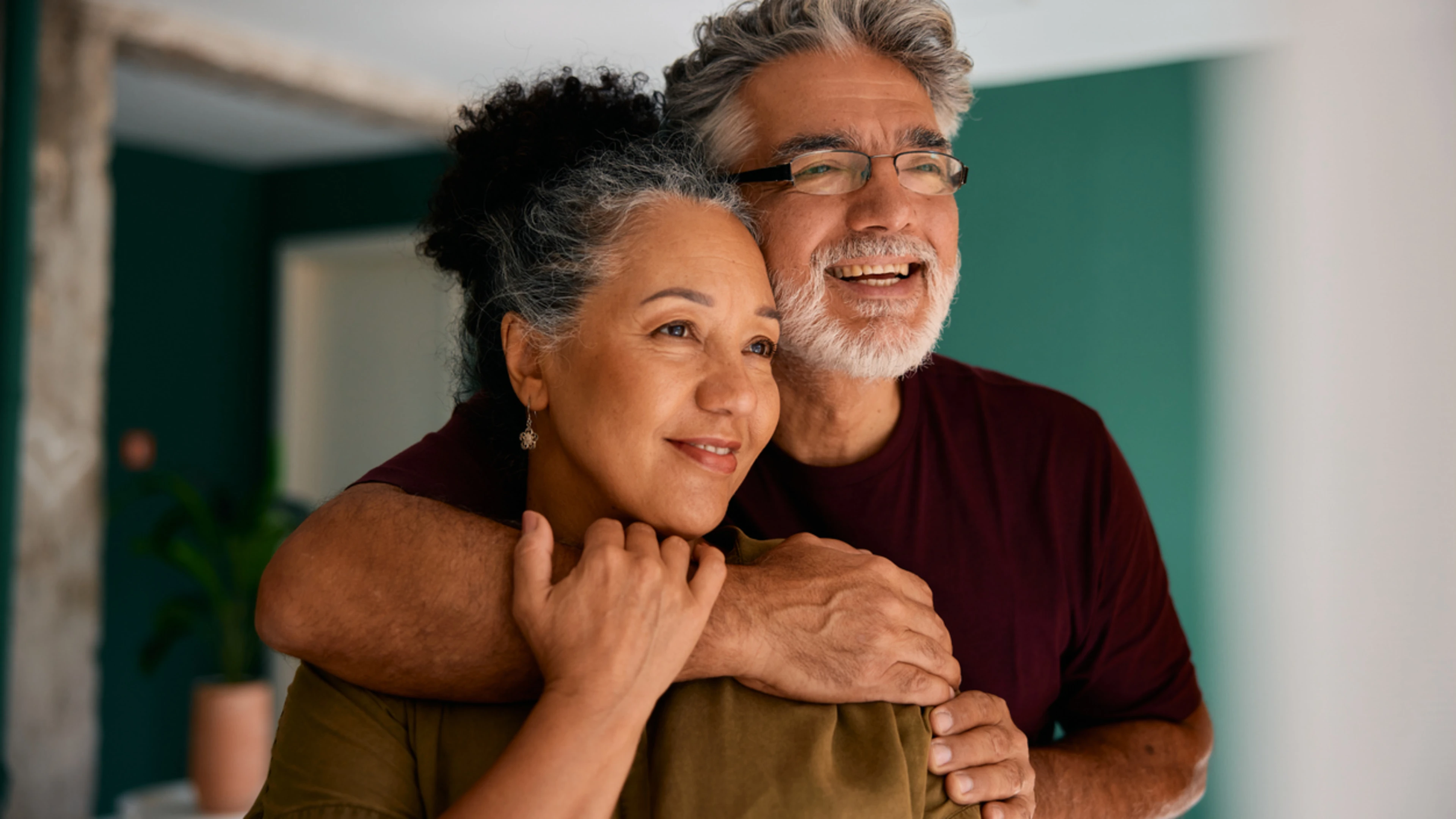 A loving couple in their 50s-60s shares an affectionate embrace in their modern home. The gray-haired man wraps his arms around the woman from behind, both laughing joyfully. A teal accent wall and natural lighting convey comfort and partnership.