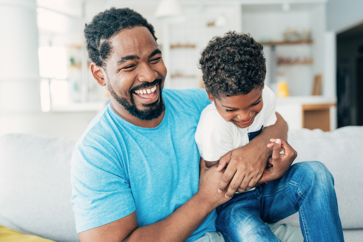 Father and son laughing joyfully on a sofa in a bright living room. The father, in a blue shirt, embraces his son, creating a warm and playful scene.