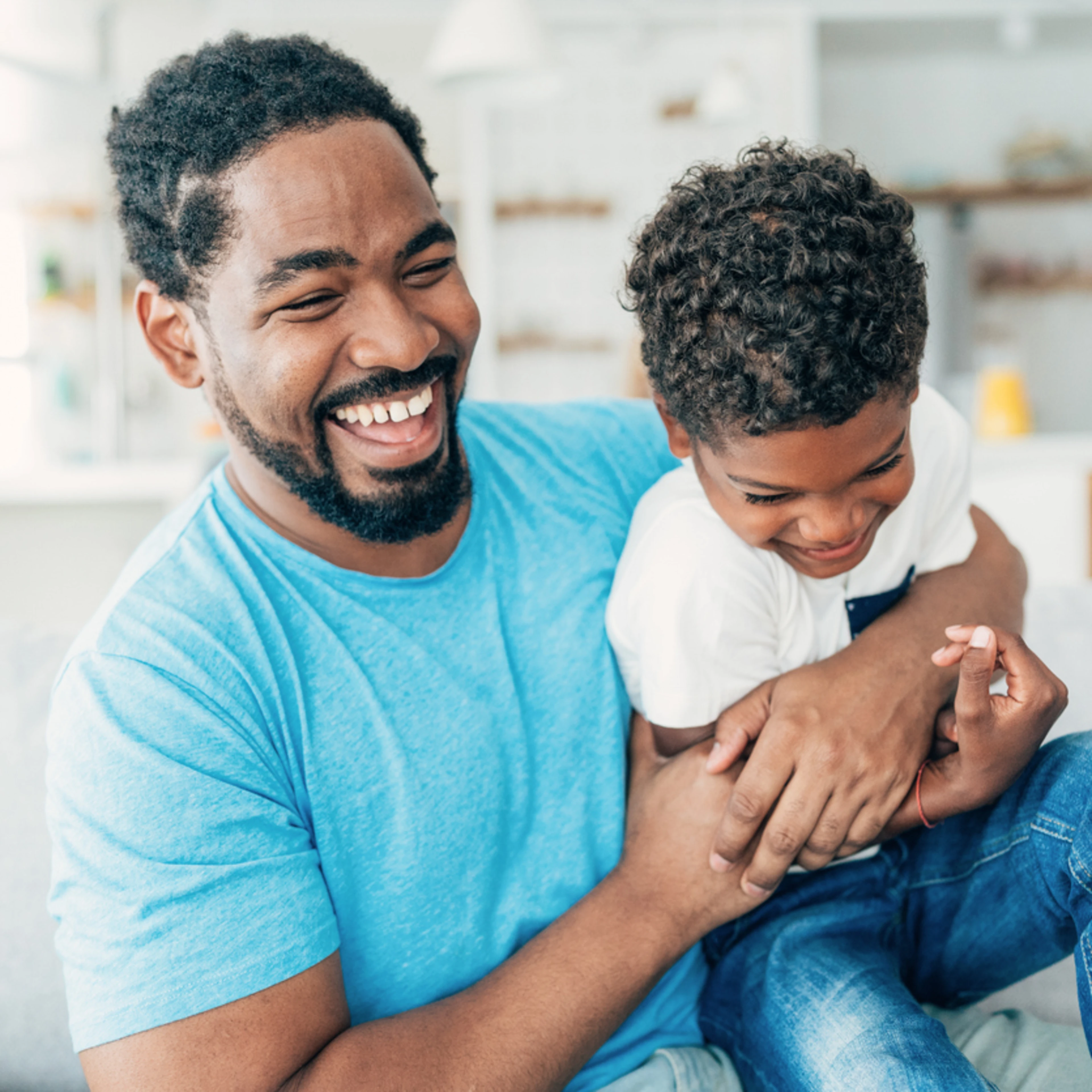 Father and son laughing joyfully on a sofa in a bright living room. The father, in a blue shirt, embraces his son, creating a warm and playful scene.