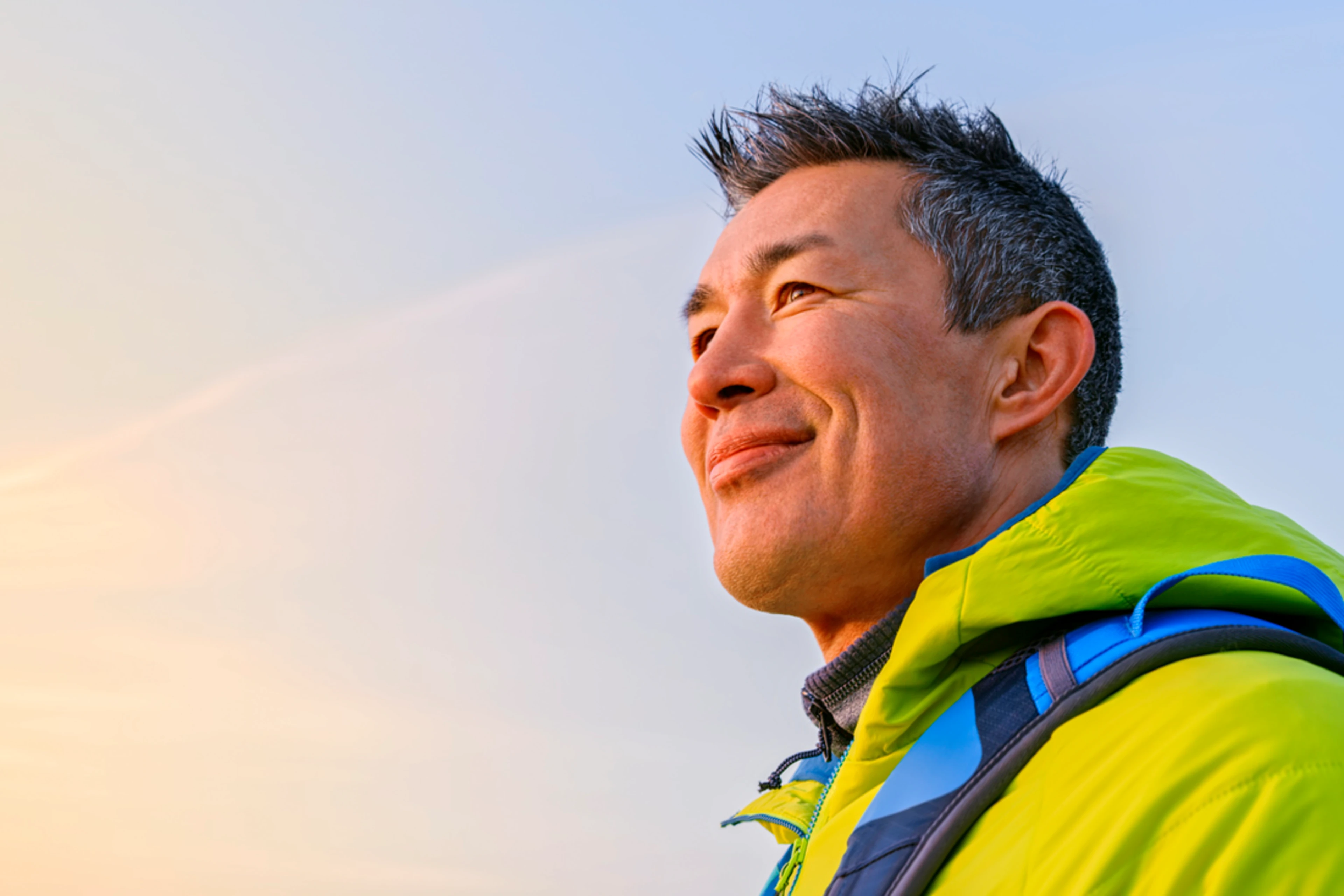 A middle-aged man with short dark hair smiles contentedly while hiking outdoors. He wears a bright lime green jacket with blue backpack straps visible. The golden hour sky creates a warm, uplifting mood of adventure and optimism.