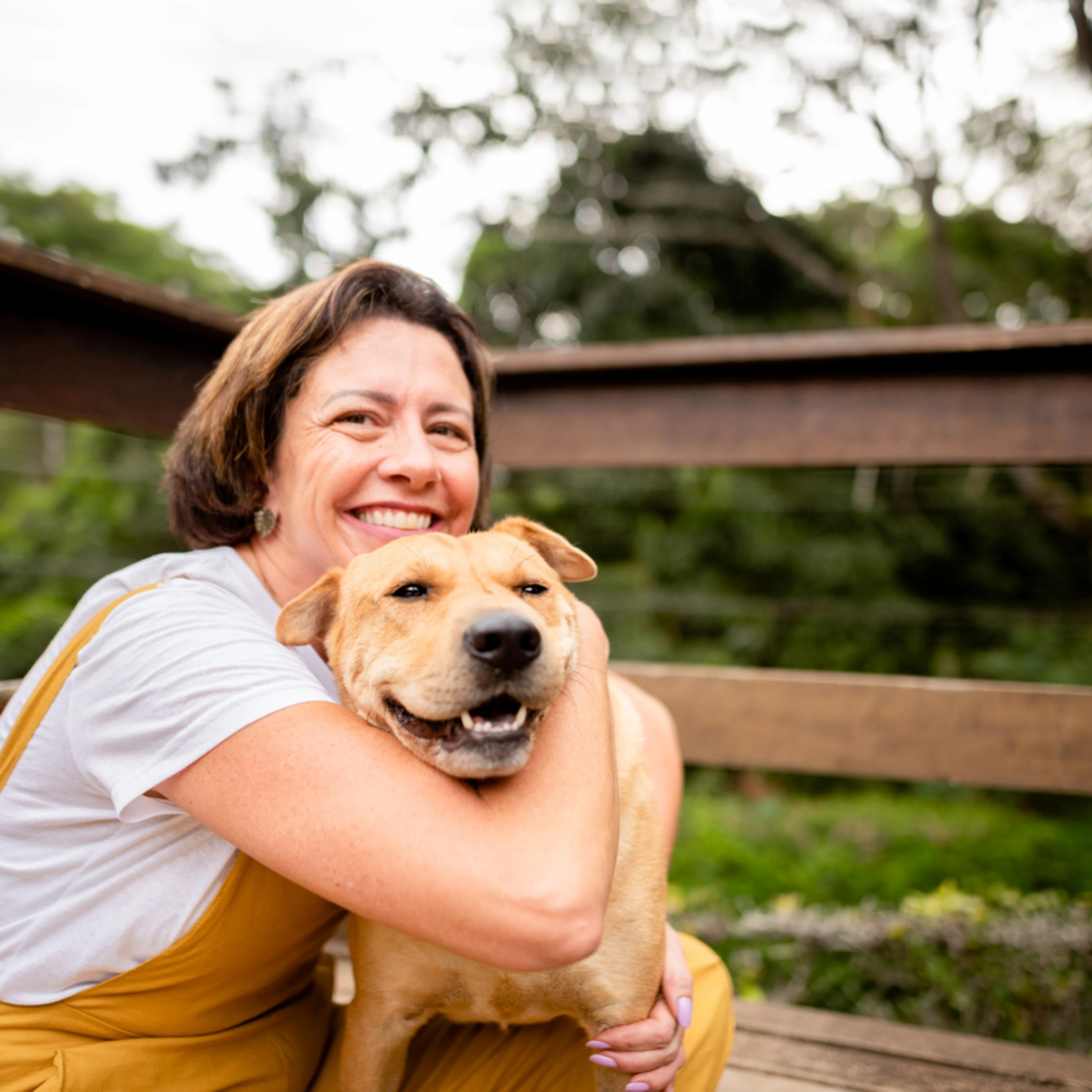 A joyful middle-aged woman with short brown hair embraces her happy tan Labrador on a wooden deck surrounded by greenery. She wears mustard yellow overalls over a white t-shirt, beaming with affection as the dog smiles contentedly.