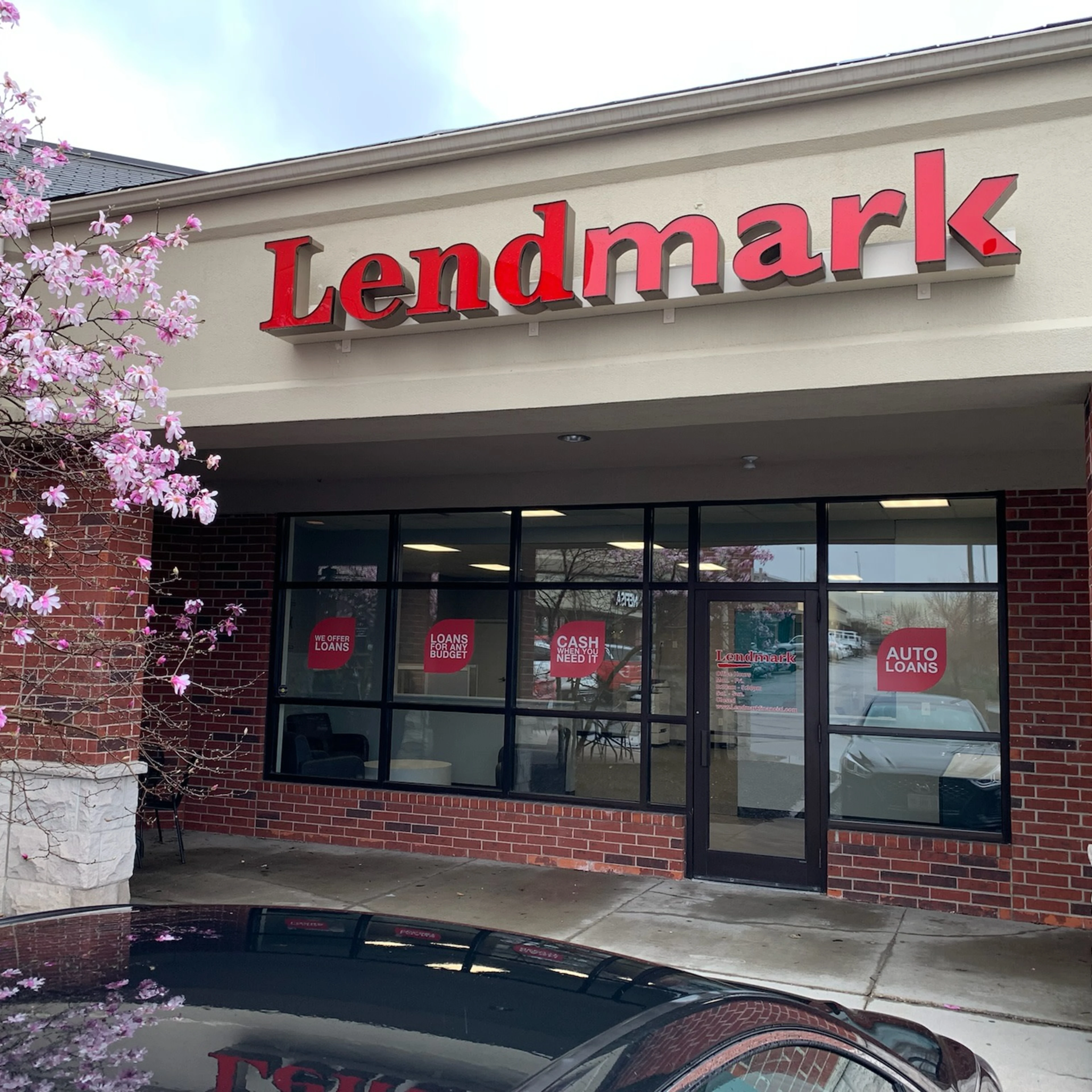 Lendmark Financial Services storefront on a spring day with red brick walls and beige stucco displaying the company name. Large windows show loan promotional signs, and a pink magnolia tree in bloom frames the welcoming entrance.