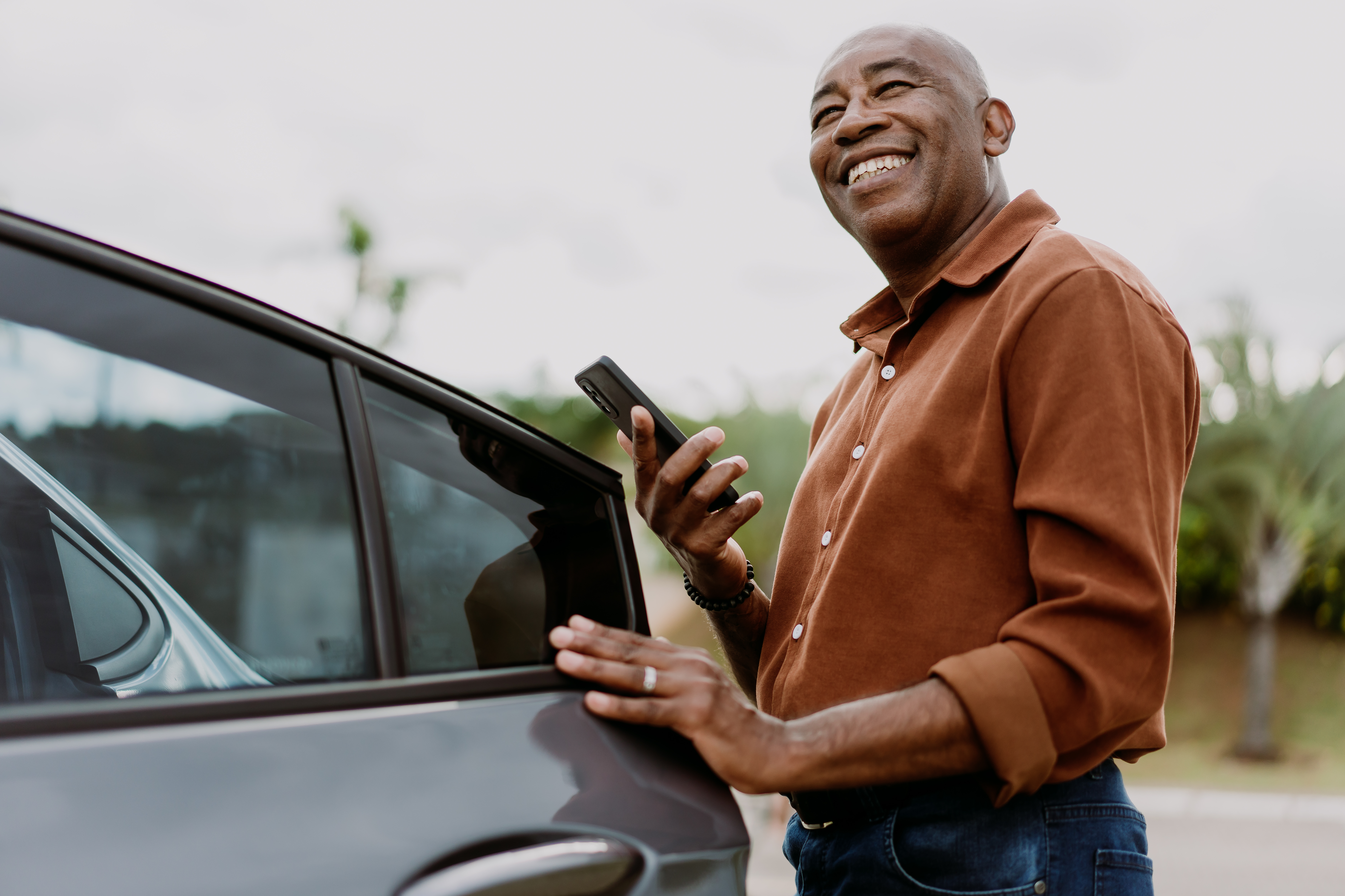 A joyful middle-aged Black man with a bald head leans against a gray sedan while holding a smartphone. He wears a rust-colored shirt and dark jeans, smiling broadly. Soft, overcast light and blurred greenery create a warm, candid atmosphere.