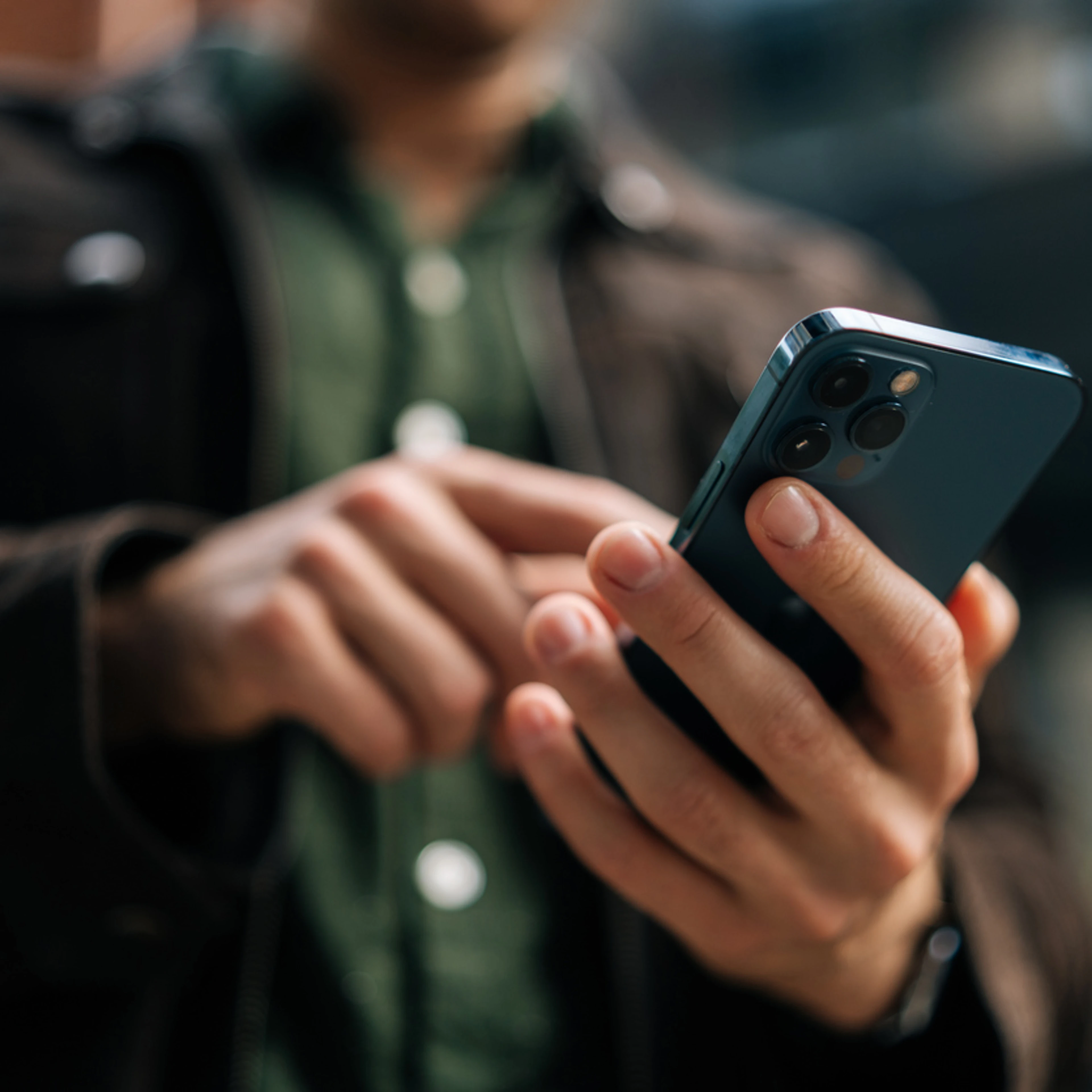 A person in a brown jacket and green shirt is using a modern smartphone outdoors. The focus is on the phone in their hand, exuding a casual, tech-savvy vibe.
