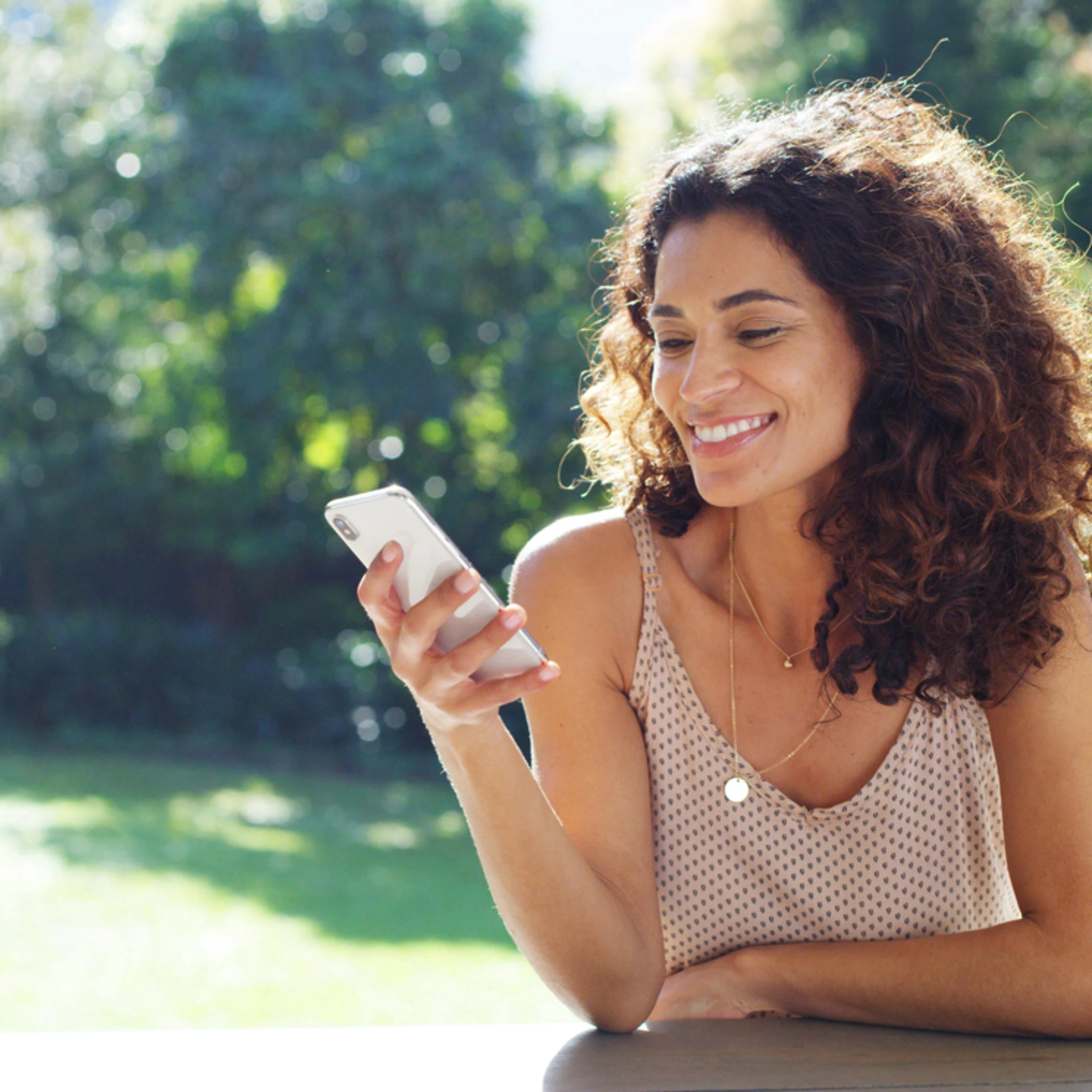 A woman with long, curly brown hair smiles warmly while looking at her smartphone on an outdoor patio. She wears a beige polka dot tank top and gold necklace. A sunlit backyard with lawn, trees, and pool creates a relaxed summer atmosphere.
