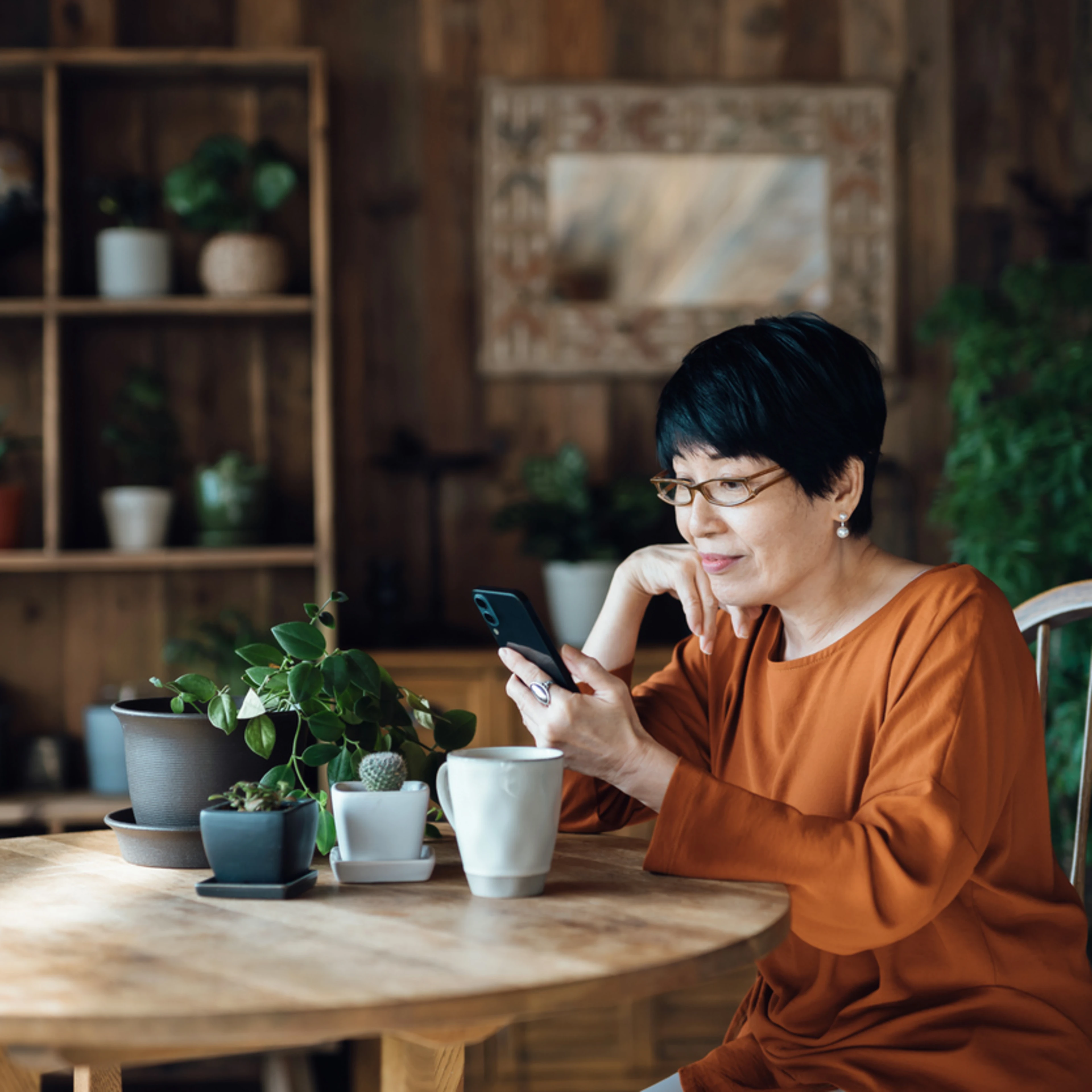A woman with short black hair and glasses sits at a table with plants and a mug, looking at her smartphone.
