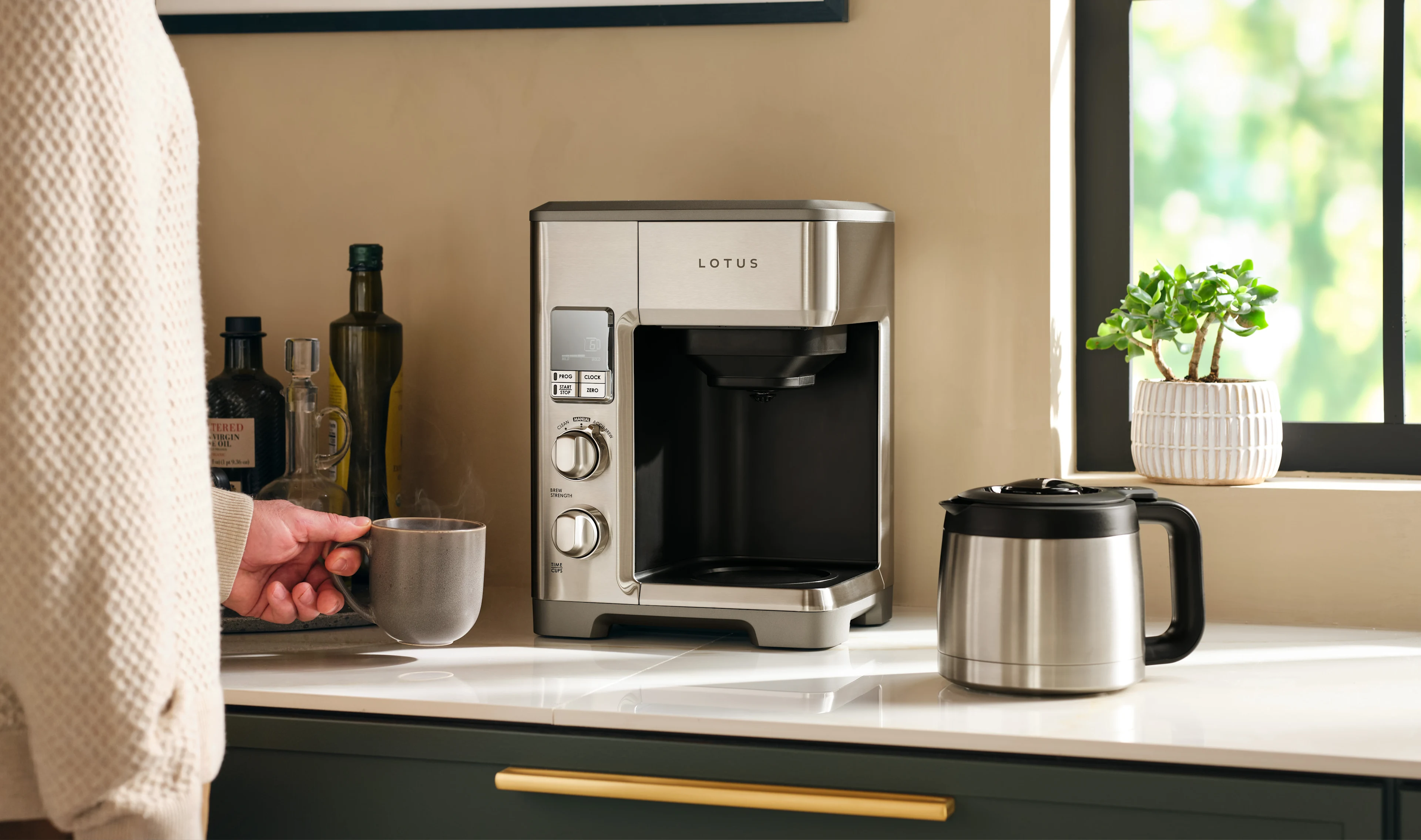 lotus stainless steel coffee maker brewing fresh coffee in a sunlit modern kitchen, with a person holding a steaming mug and thermal carafe nearby