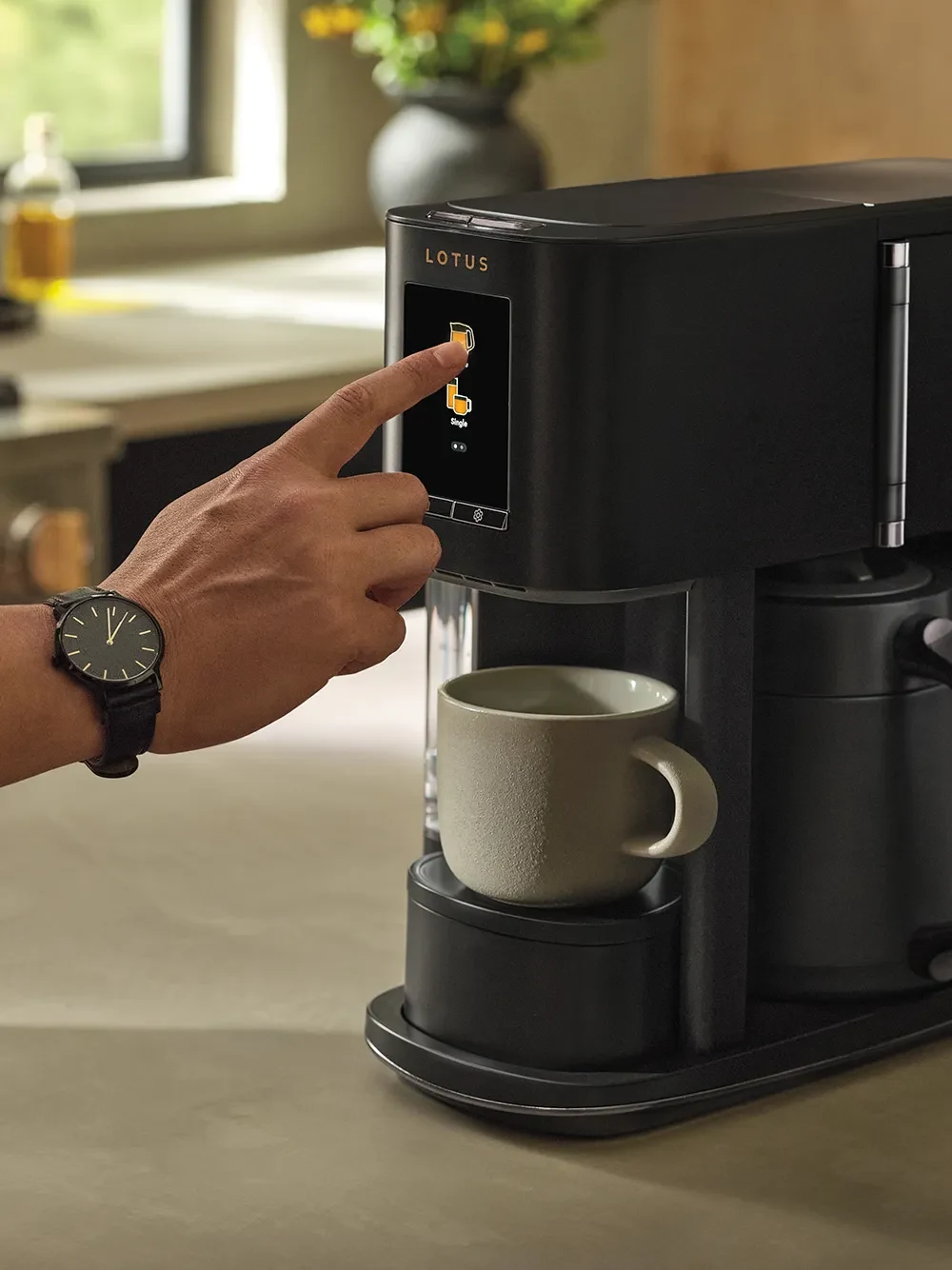 The Ritual coffee system in Obsidian with hand selecting a brew option on the touchscreen, and a mug beneath the dispenser