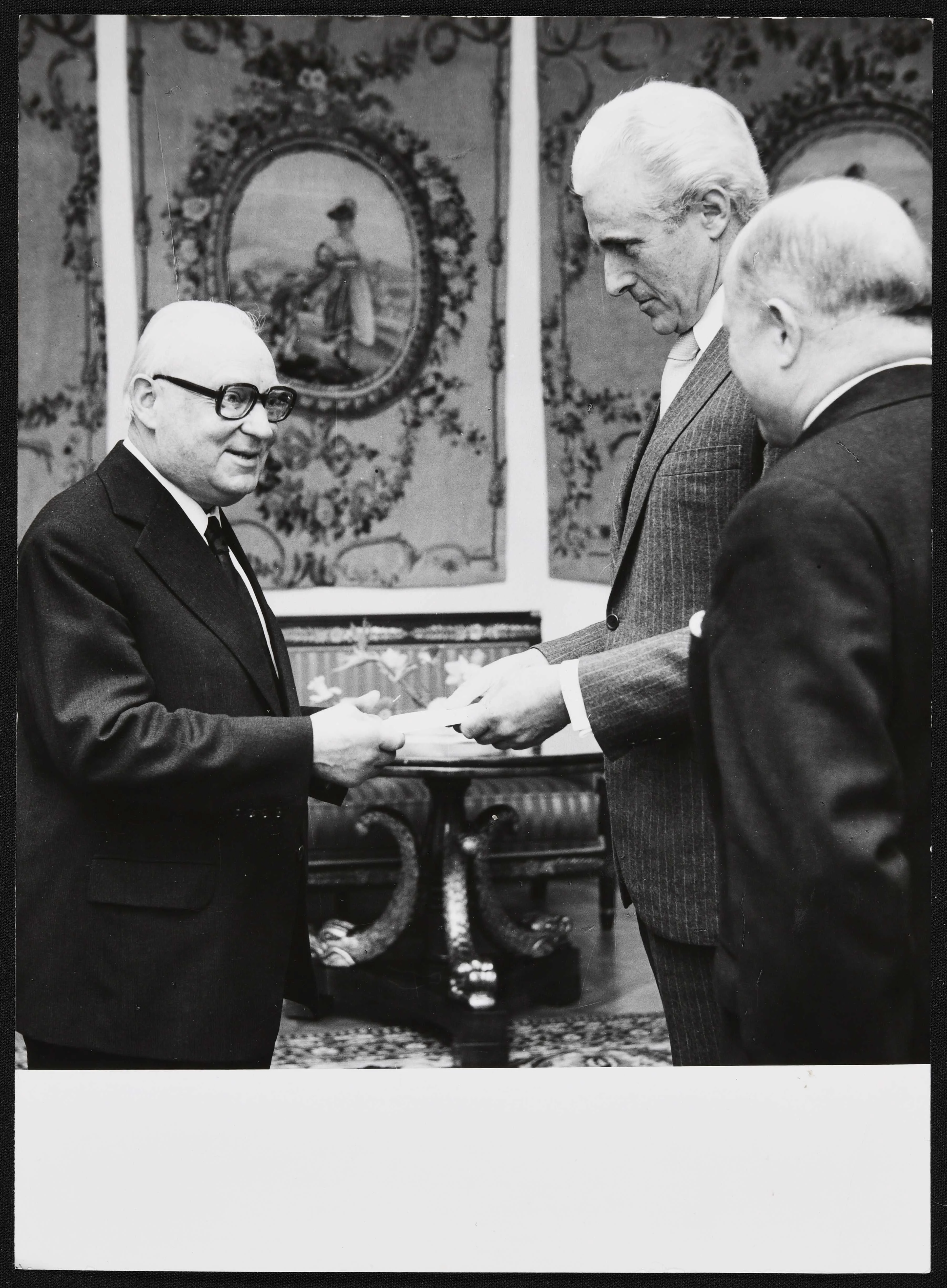 2 men exchange documents while another looks on.