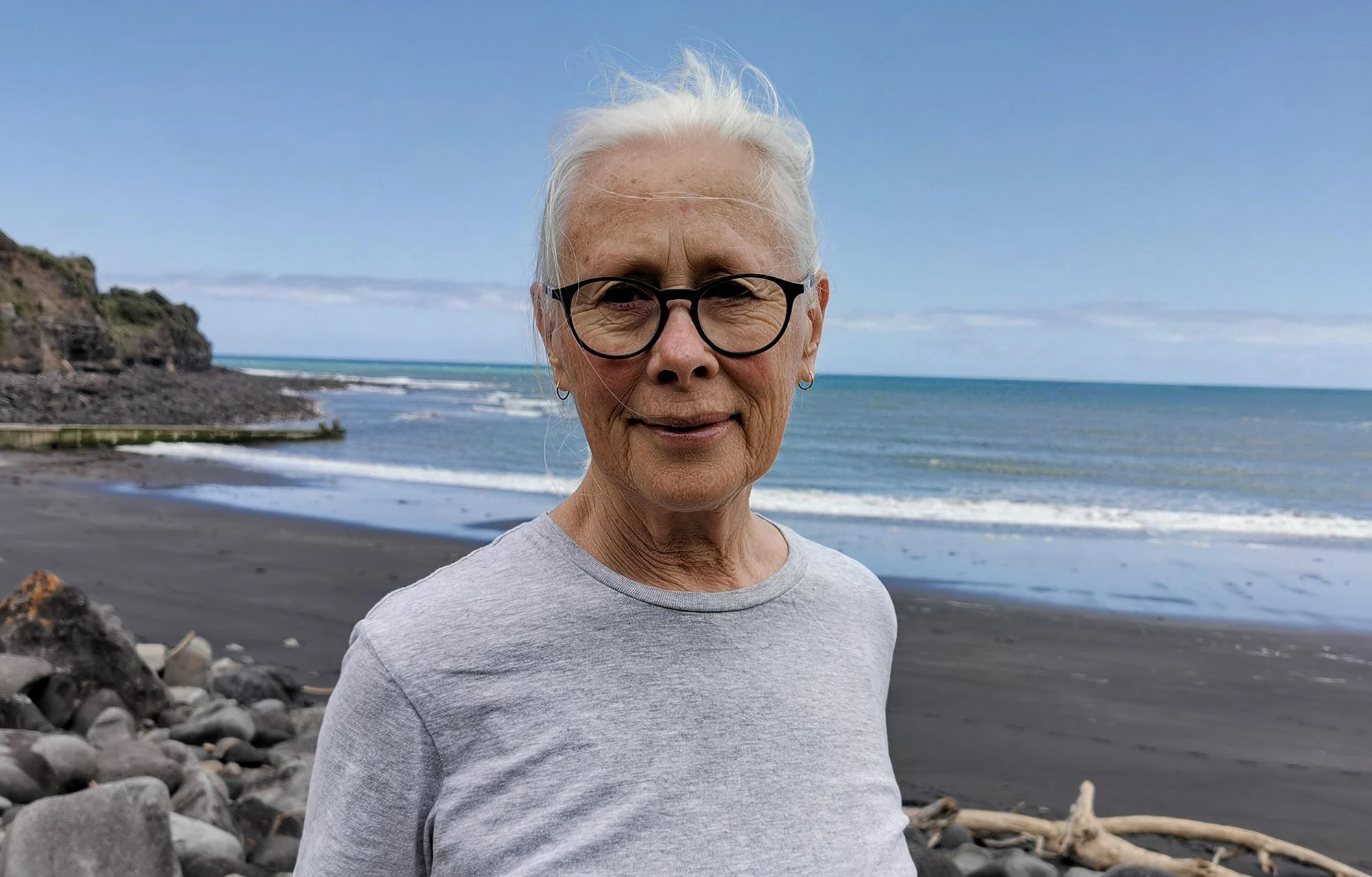 Old woman with white hair wearing glasses and a grey t-shirt, standing on a beach