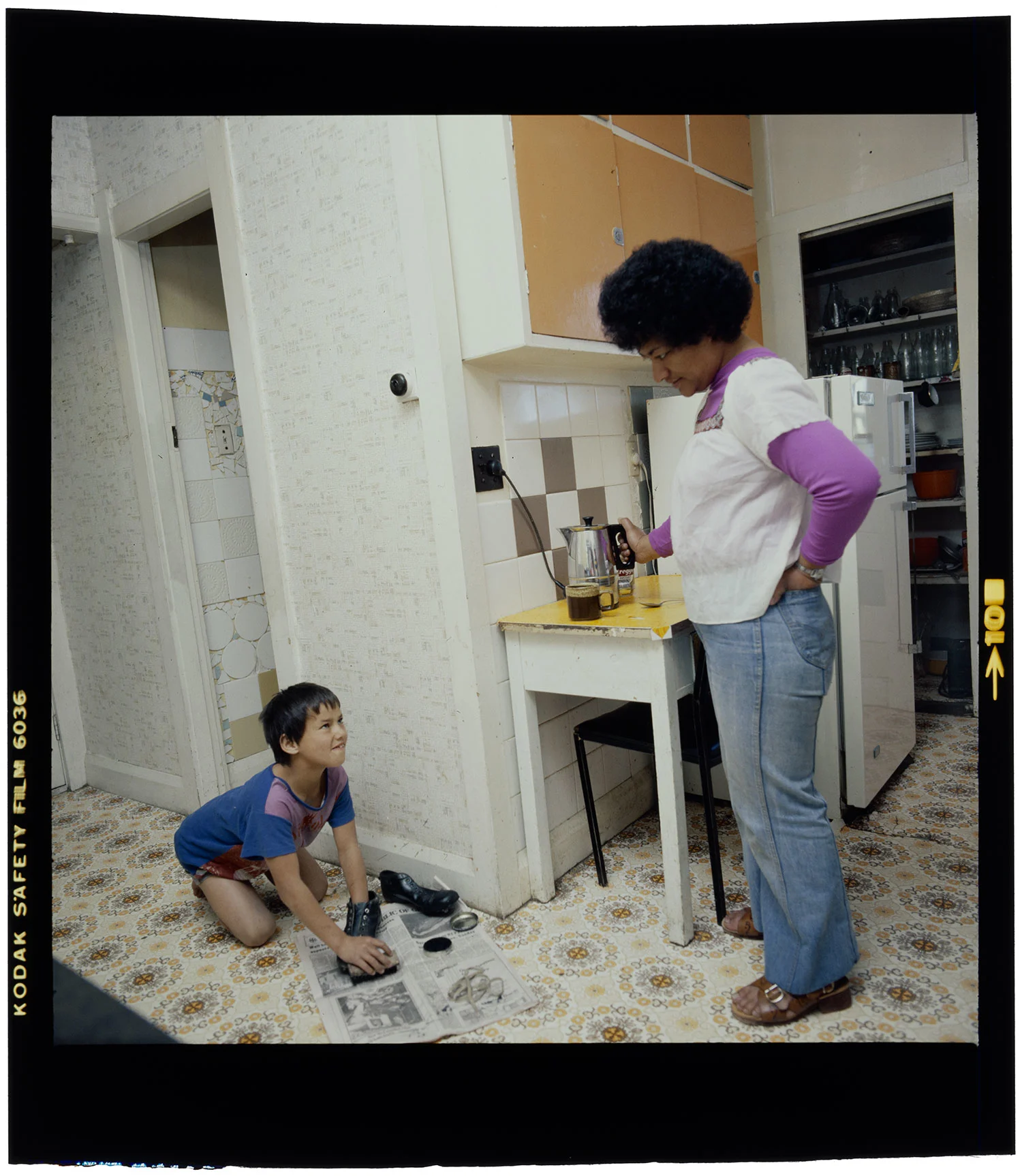 A boy in blue shirt sitting on a kitchen floor polishing his shoes while a woman with short hair wearing a purple full sleeves shirt and white top standing in front of him