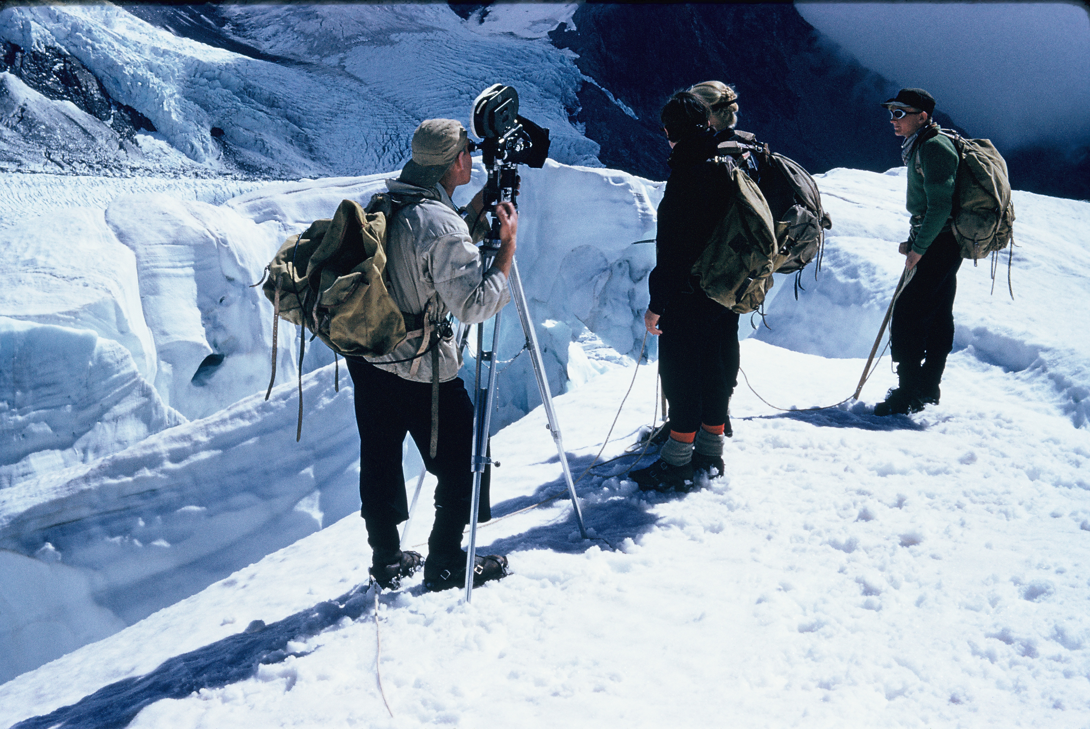 3 mean standing on snow with mountaineering gear. Man on the left is standing behind a film camera