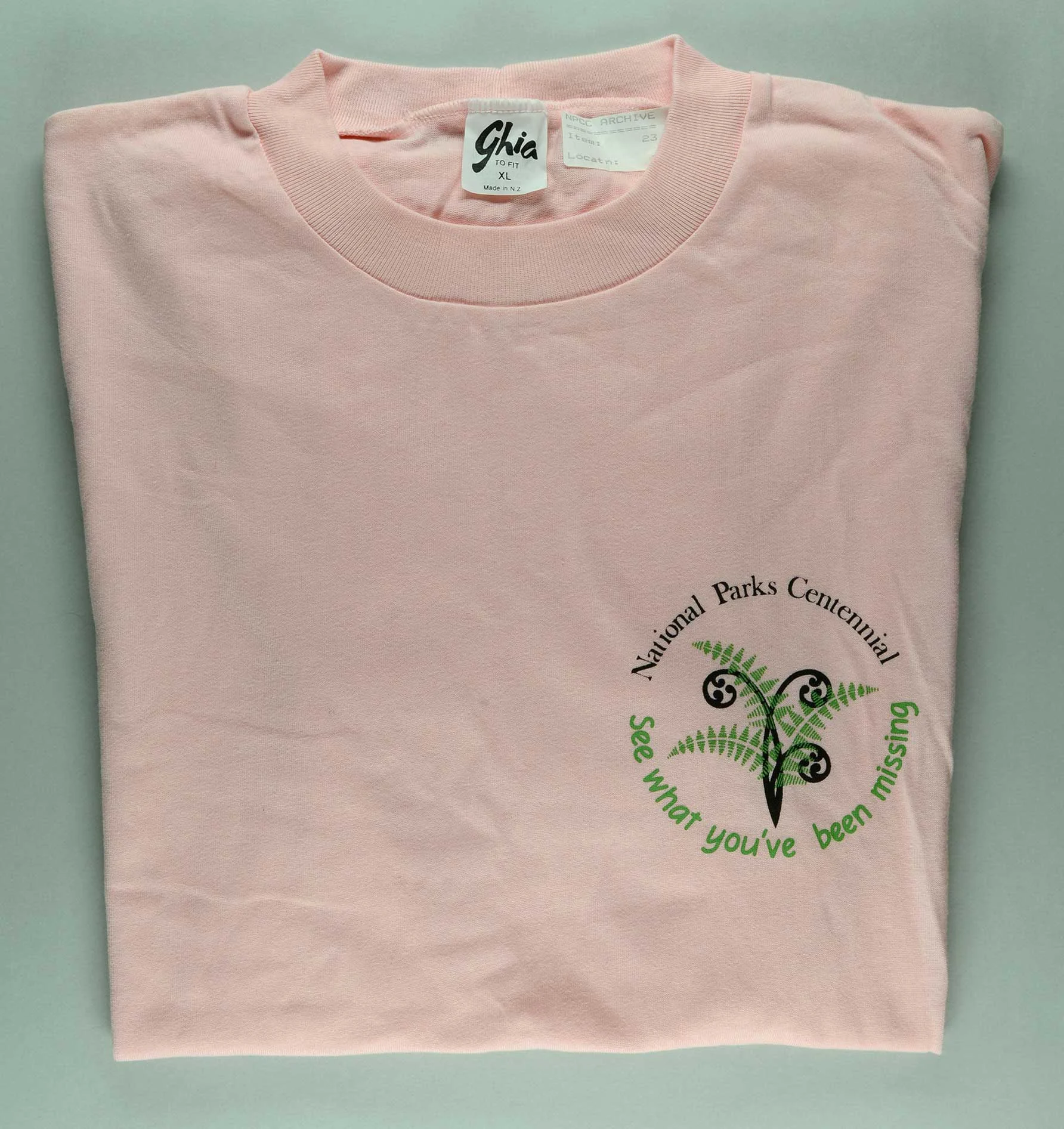 A folded pink T-shirt with a green fern and black koru and come text written in a circle around then logo.  