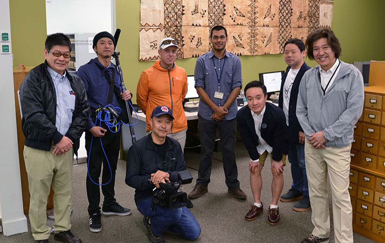 Seven people smiling for a photo and one person sitting holding a camera with a painted bark cloth on the wall behind