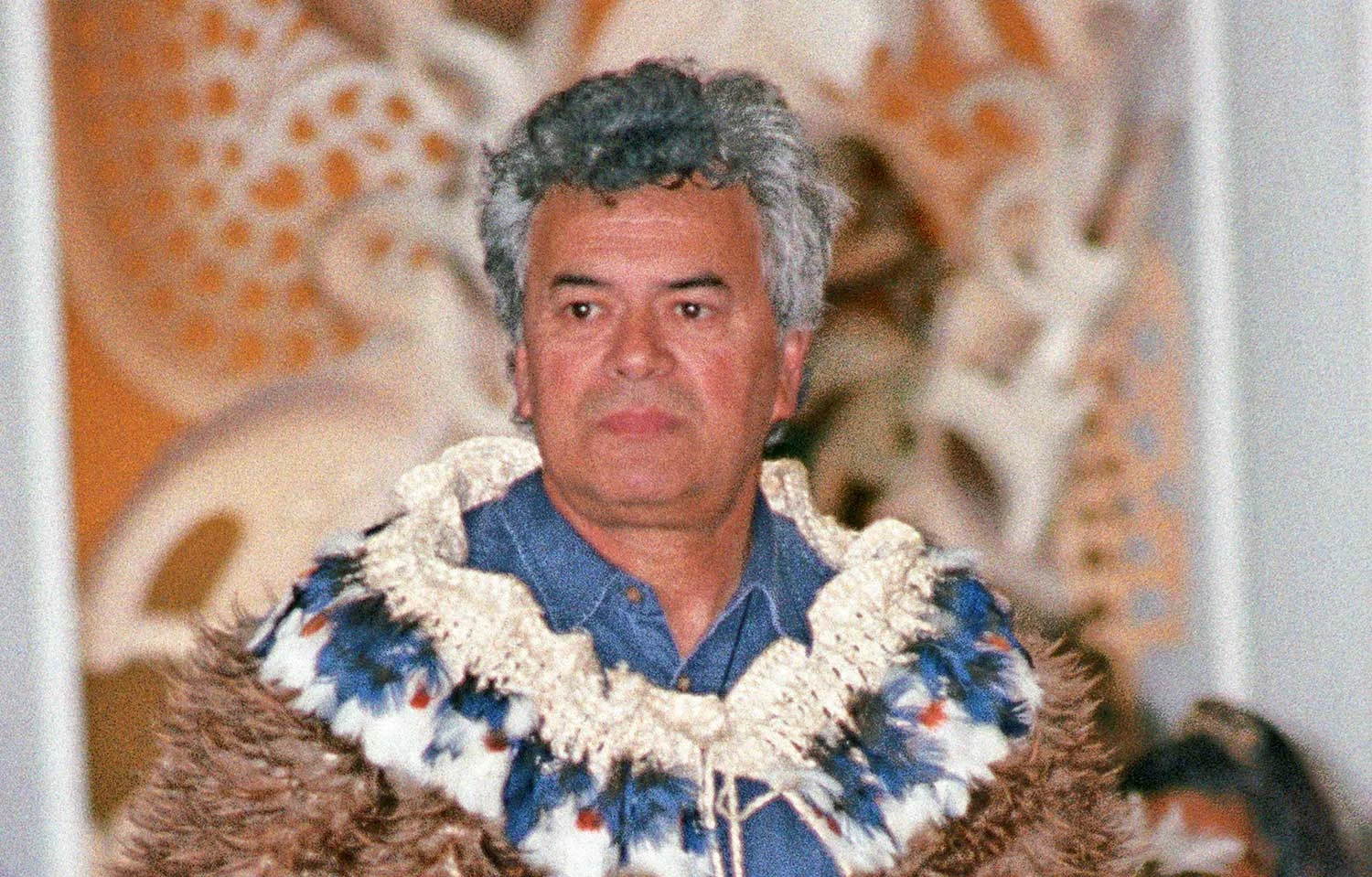 A man with grey hair wearing a Māori cloak standing in front of a colorful painting