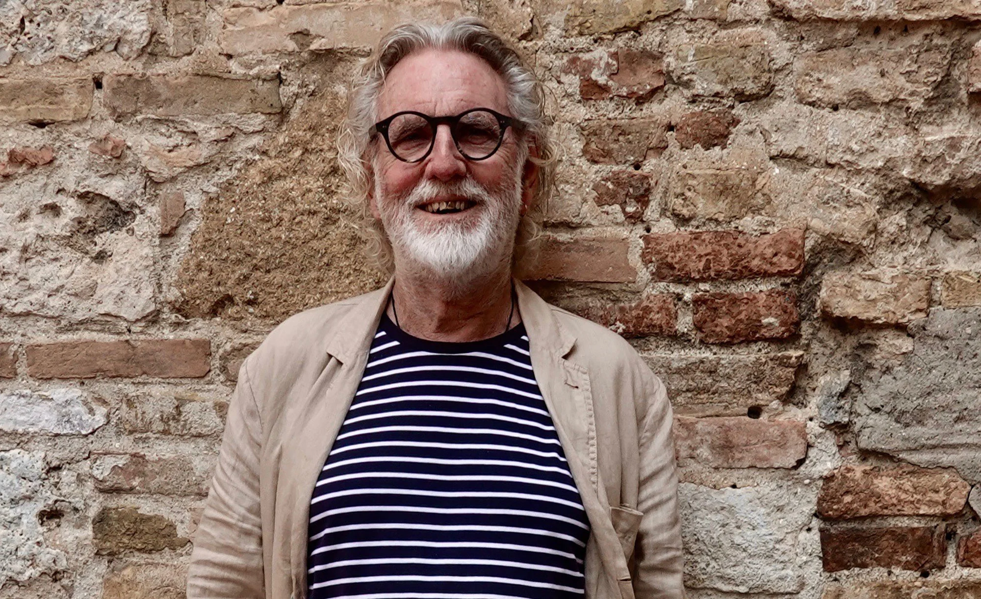 An smiling old man with grey hair and beard wearing a striped shirt and khaki jacket, standing against a brick wall.