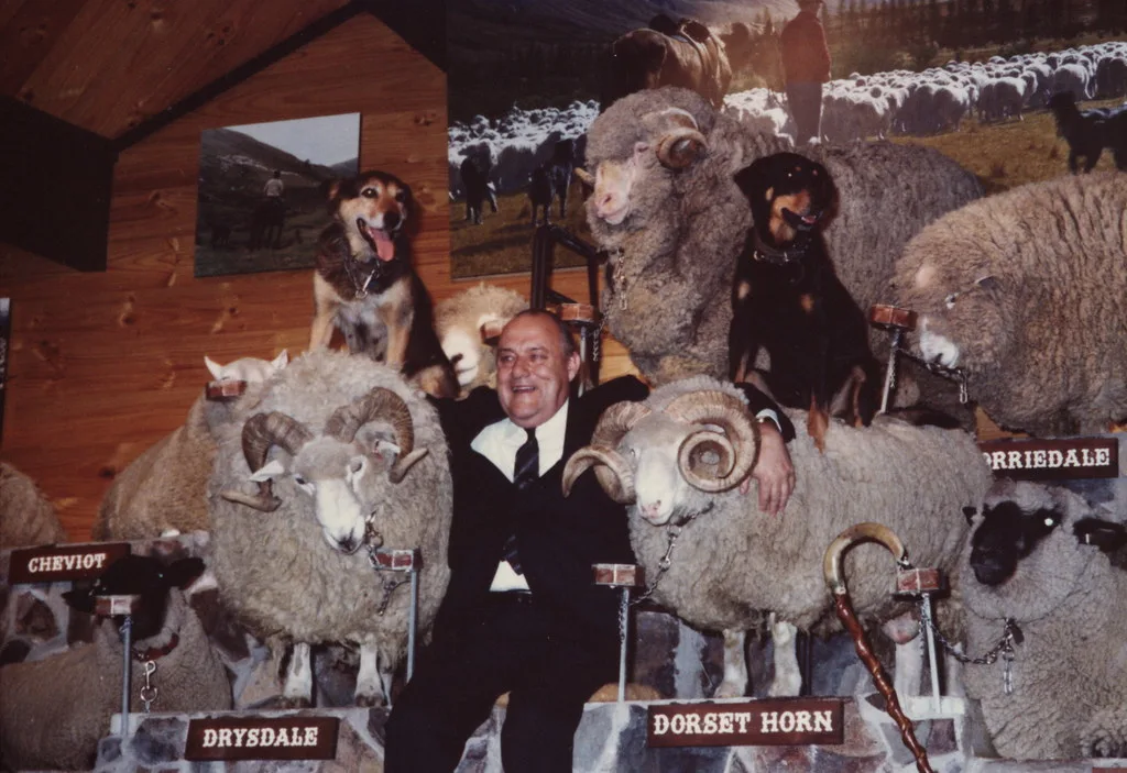 man in a suit sitting on a stage with his arms around two sheep with two dogs sitting on the sheeps backs