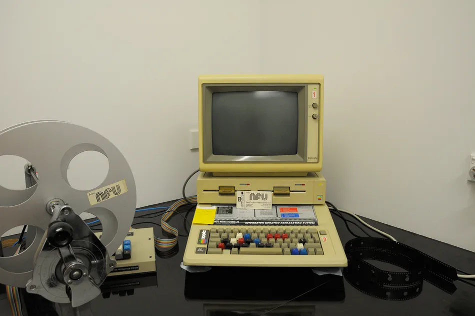 An Apple IIe computer sitting on a table. It has yellowed with age. To the left of the image is a film reel labelled with the NFU logo.