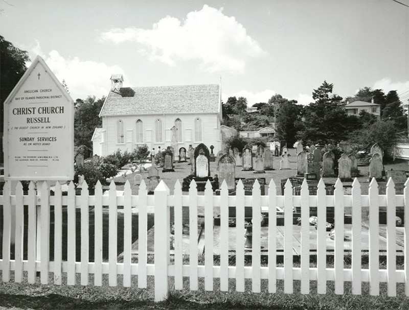 Black and white photo of a church with white fence in the front and sign board on the left