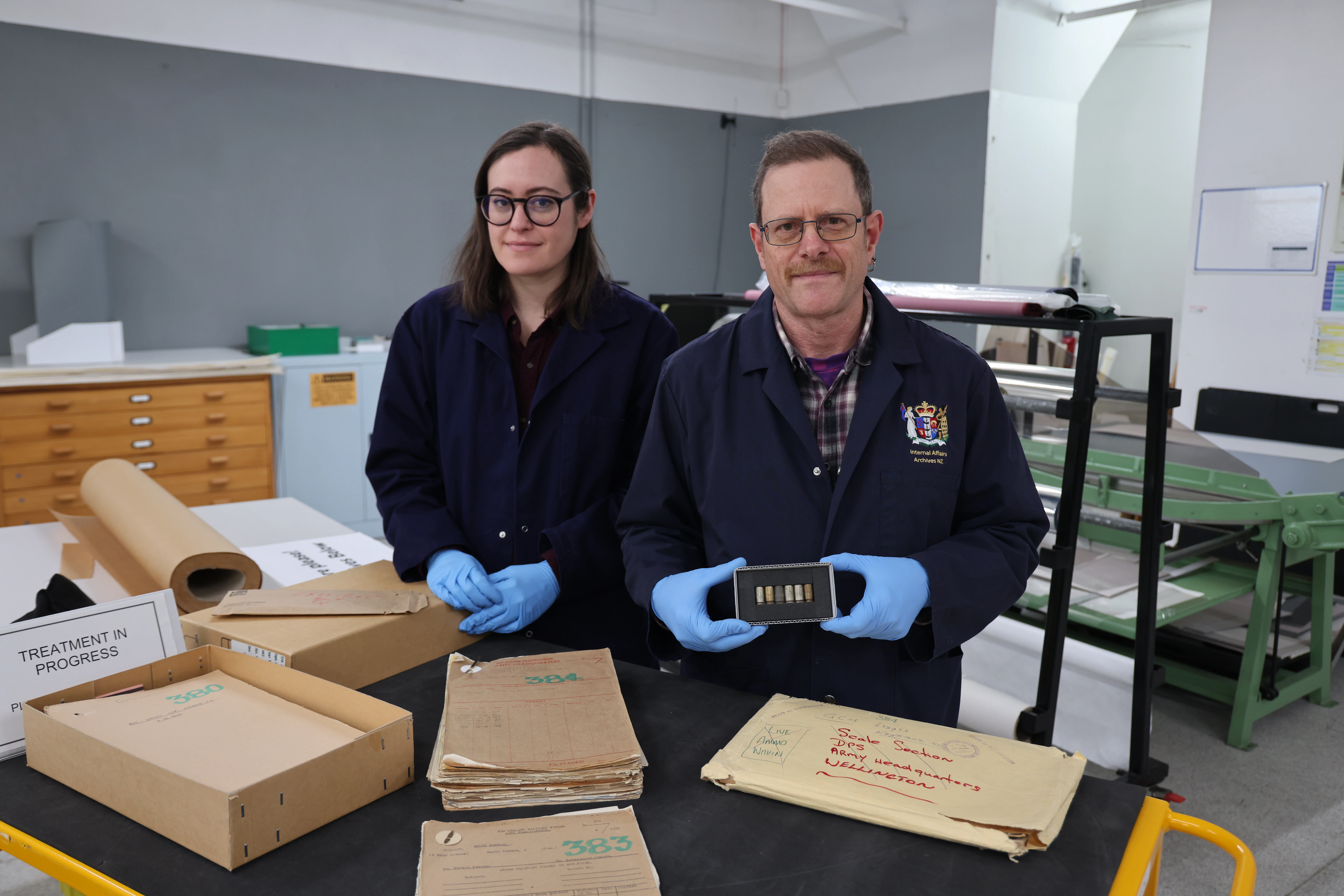 2 individuals in navy blue lab coats and light blue gloves standing at a table in a conservation lab, holding an artifact, with documents and labelled boxes marked 'Treatment in Progress' around them, suggesting a document preservation setting.