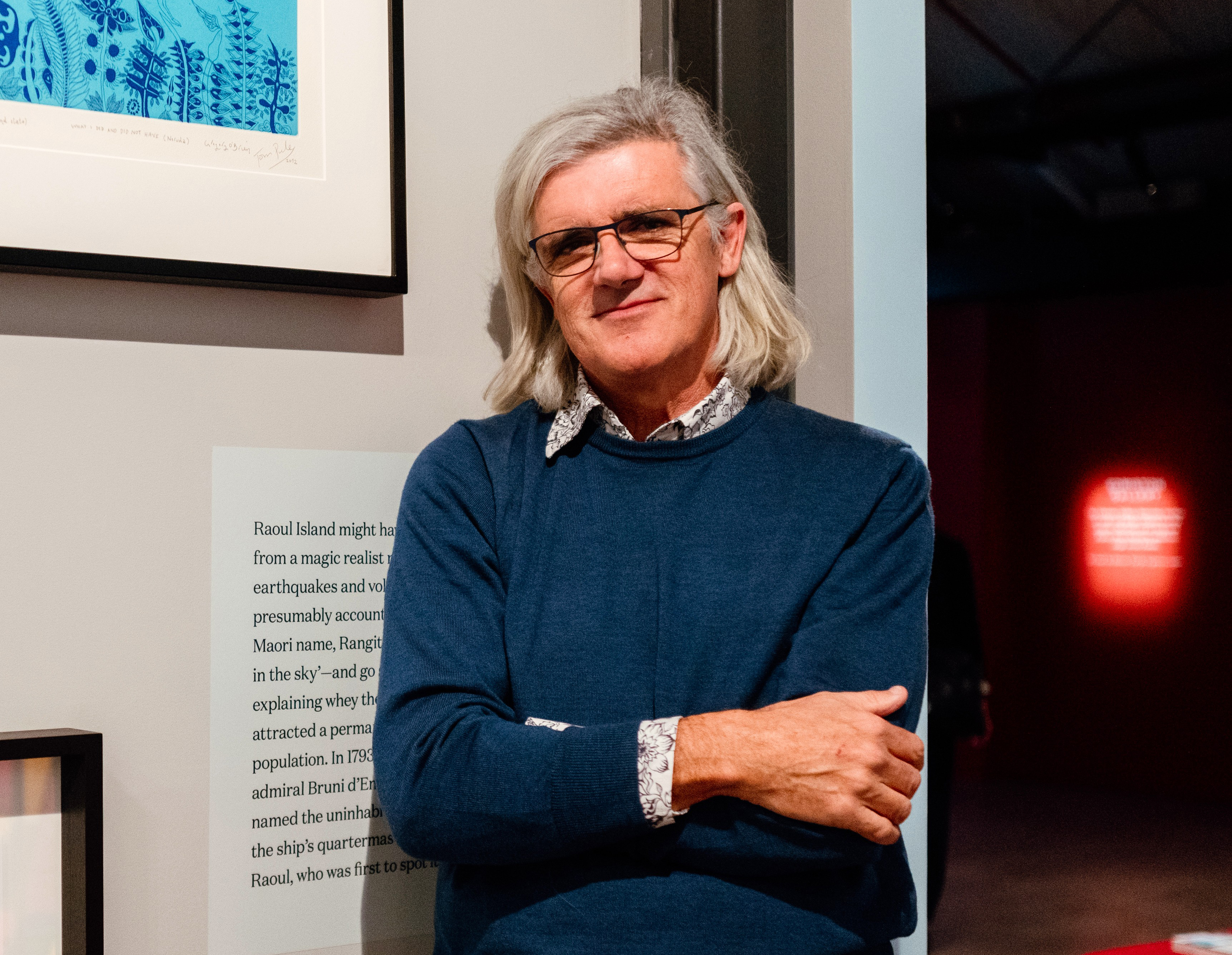 Image of Greg O'Brien in a blue jersey with a white shirt underneath, standing by one of his artworks with crossed arms.