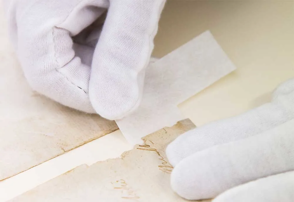 Two hands wearing white gloves sticking a tape to an old paper