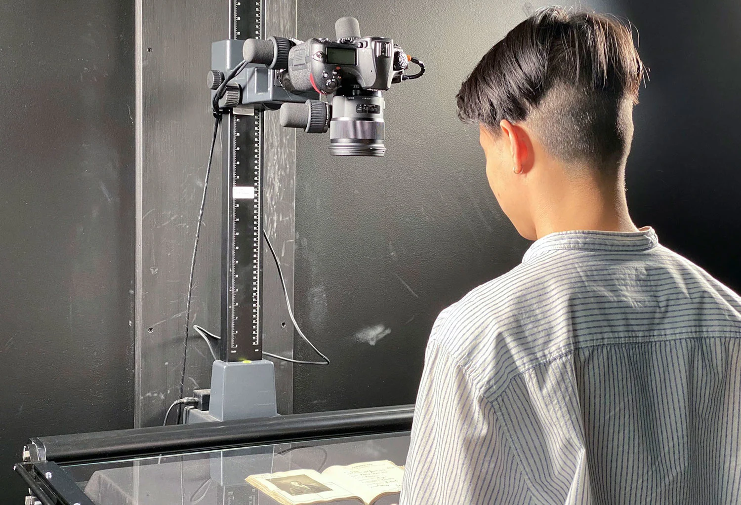 A young man with short hair facing his back to the camera and standing in front of a camera and a book laid on the glass