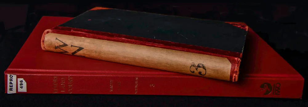 Image of reproduction copy of a Māori Land Court Minute Book lays flat with its spine to the viewer. Resting on top of the reproduction copy at a thirty degree angle is the original version of the Minute Book. Both books are Wellington minute book number 3