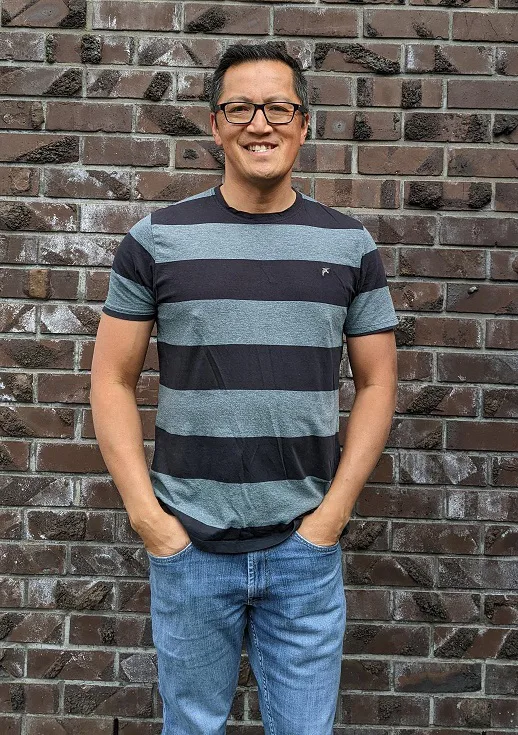 A man wearing glasses, striped blue T-shirt and blue jeans standing in front of a brick wall