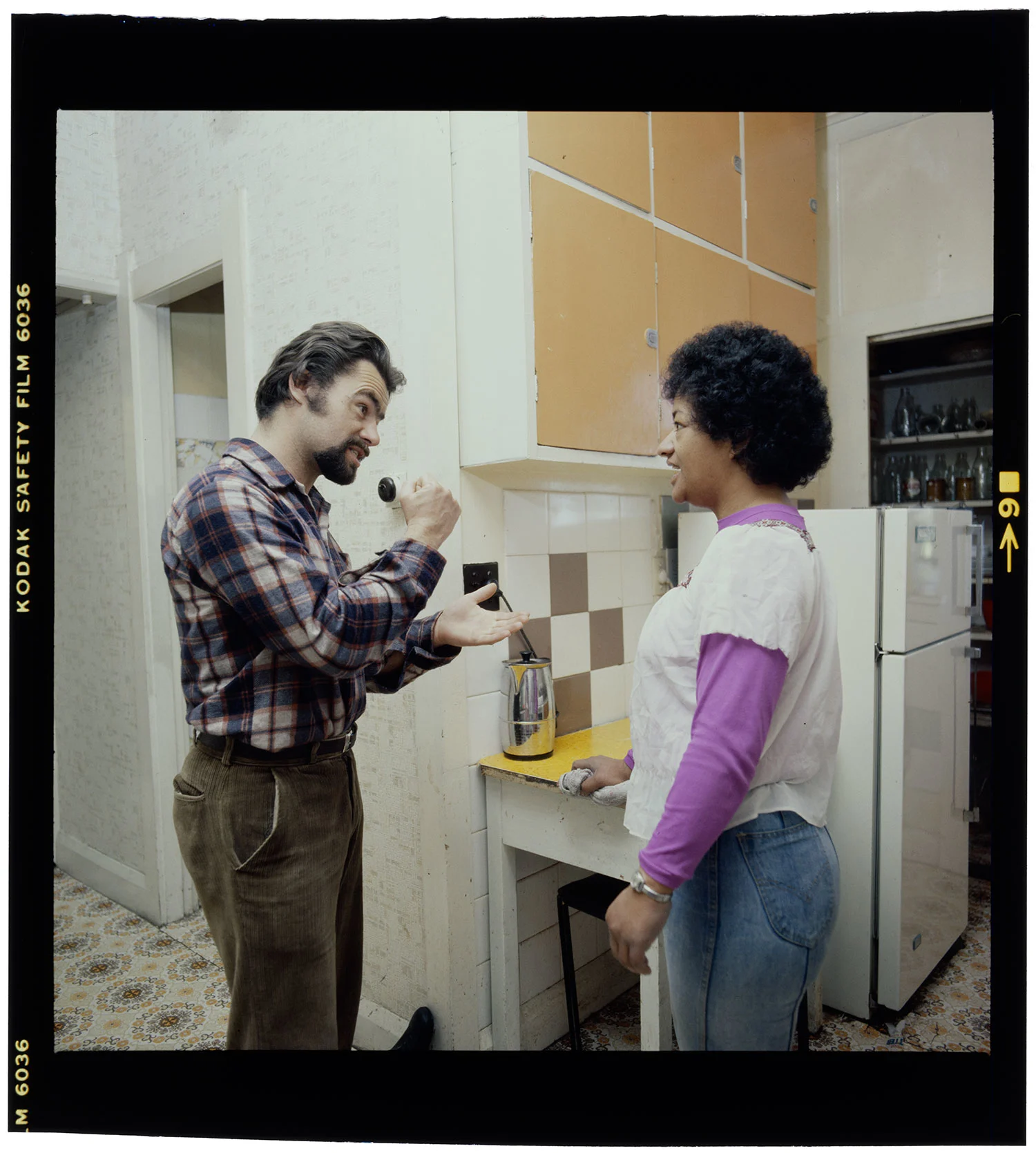 A woman with short hair wearing a full sleeves purple shirt and white top standing in a kitchen talking to a bearded man wearing a checkered shirt